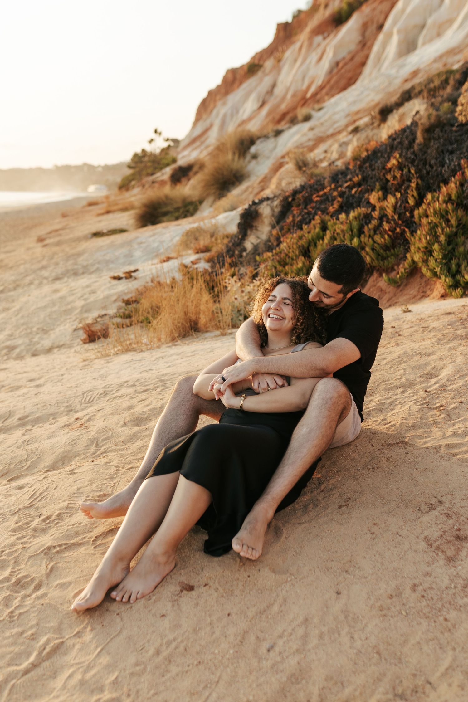 A couple embraces while sitting on a sandy beach at sunset with cliffs in the background.