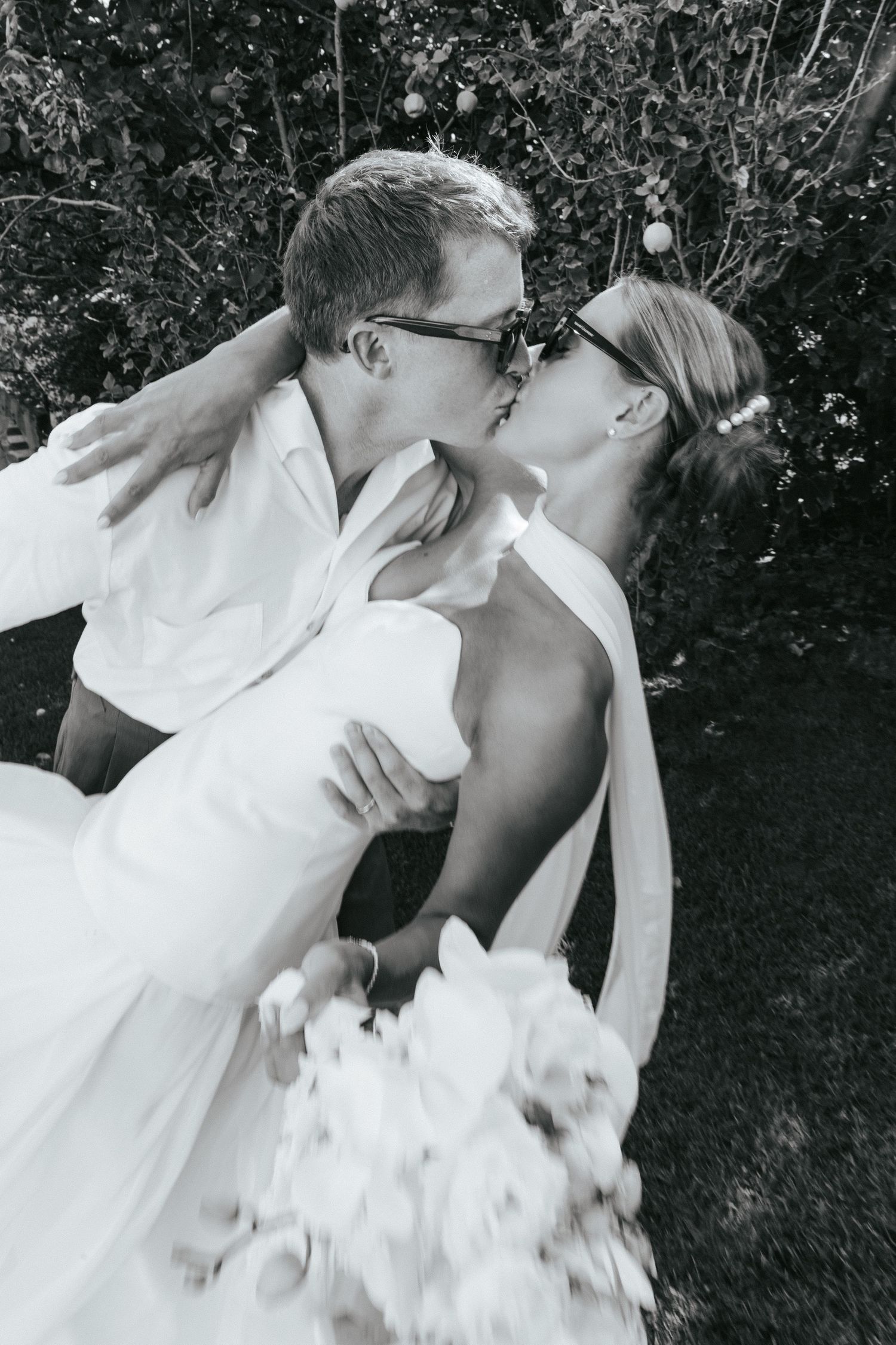 Black and white photo of bride and groom kissing in a romantic dip pose, both wearing sunglasses.