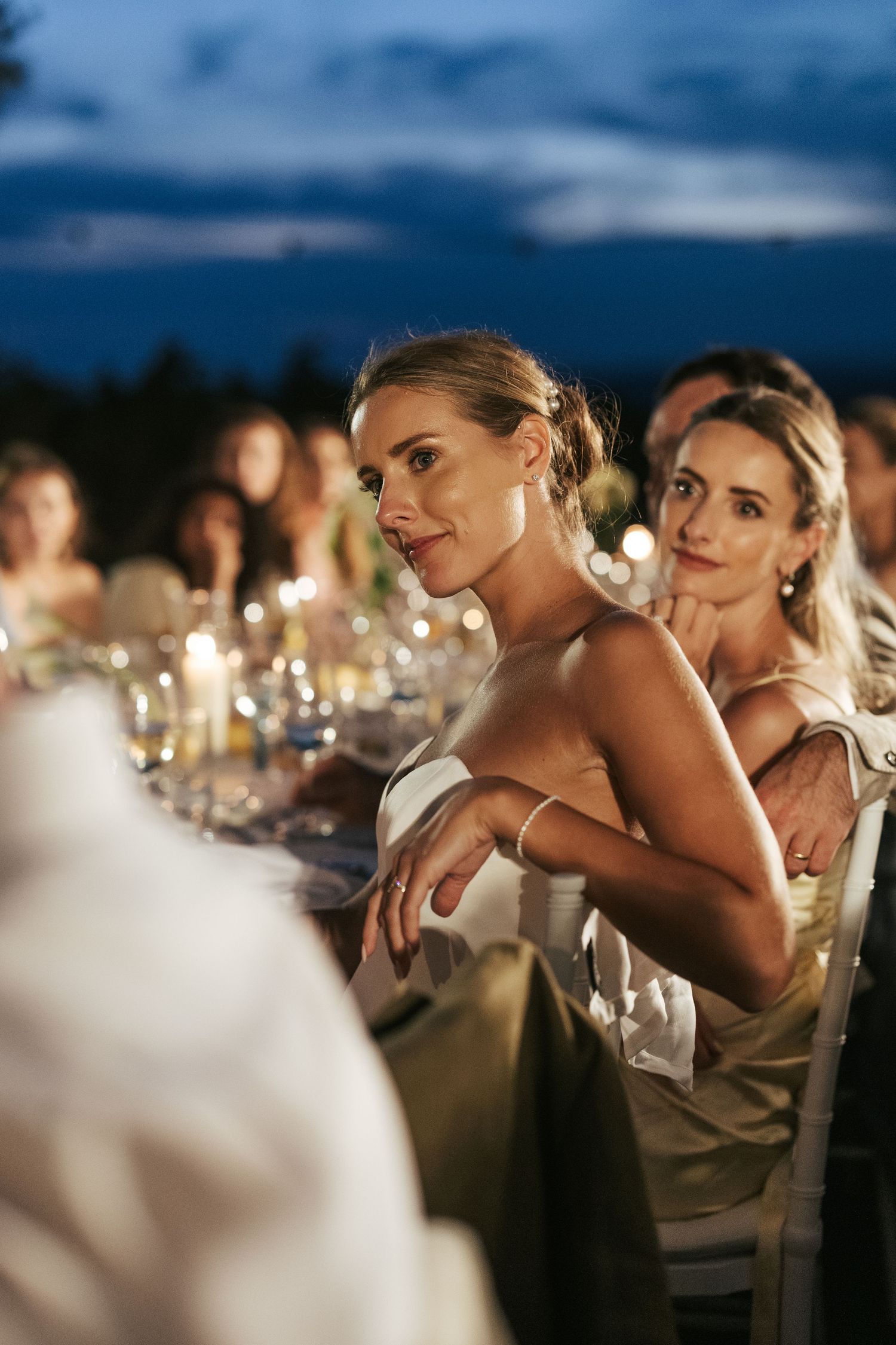 Elegant blonde woman in white dress listens attentively at candlelit outdoor evening wedding dinner.