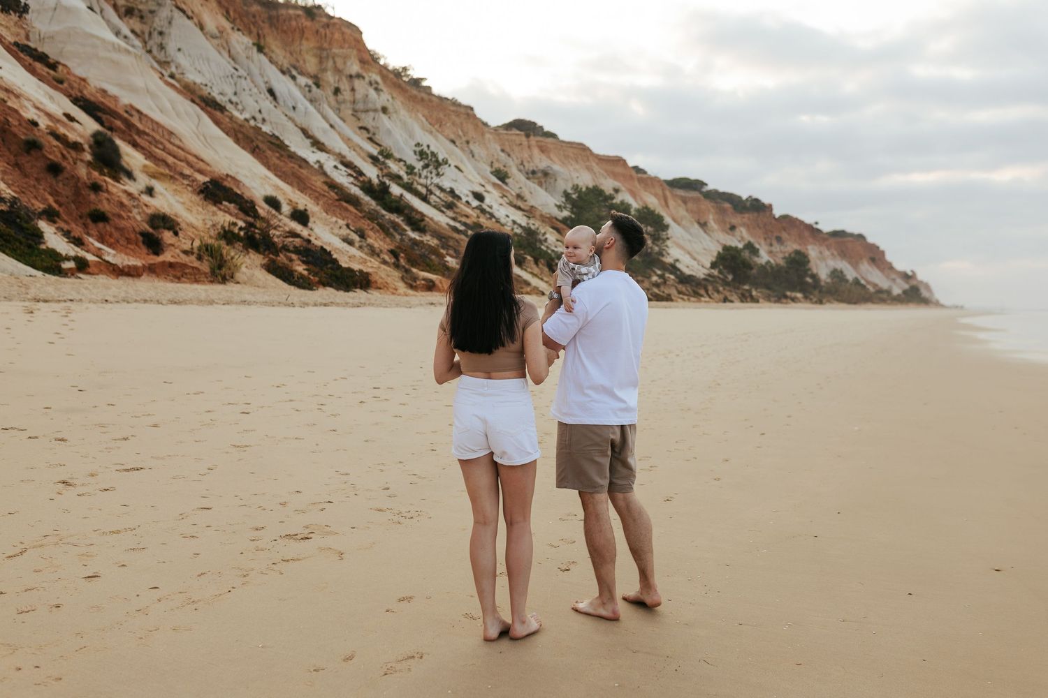 A couple in white outfits stands on a sandy beach with red sandstone cliffs rising dramatically in the background.