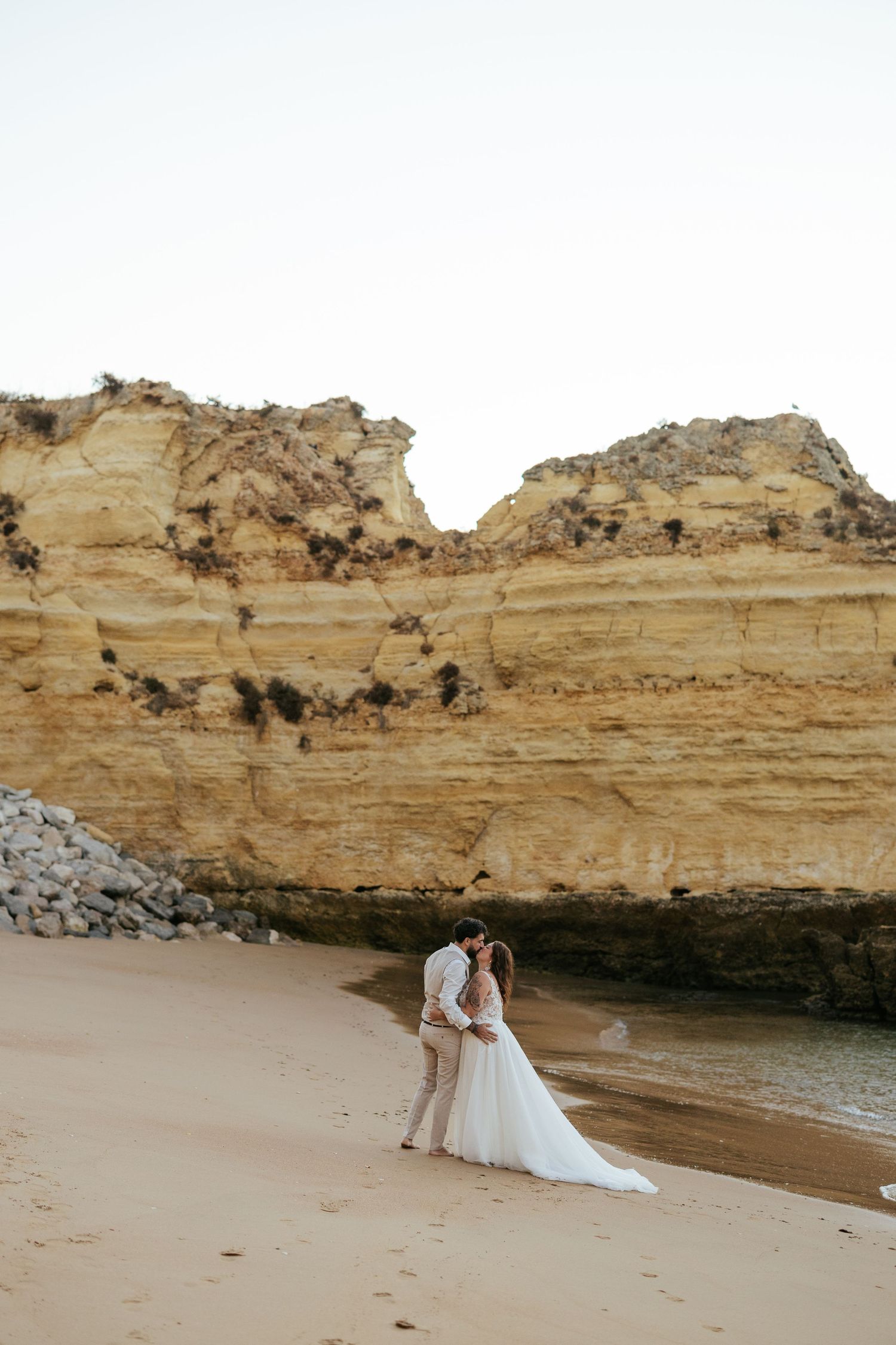 Wedding couple on sandy beach with towering limestone cliff wall backdrop. Praia Dona Ana, Lagos, Algarve, Portugal.