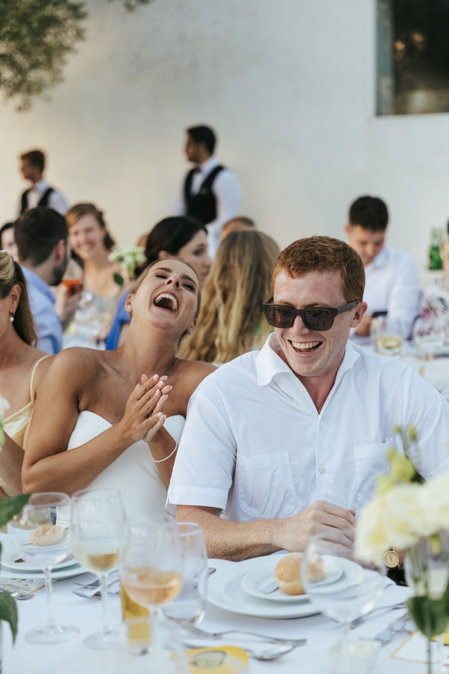 Bride and groom laughing joyfully together at wedding reception table surrounded by guests and floral arrangements.