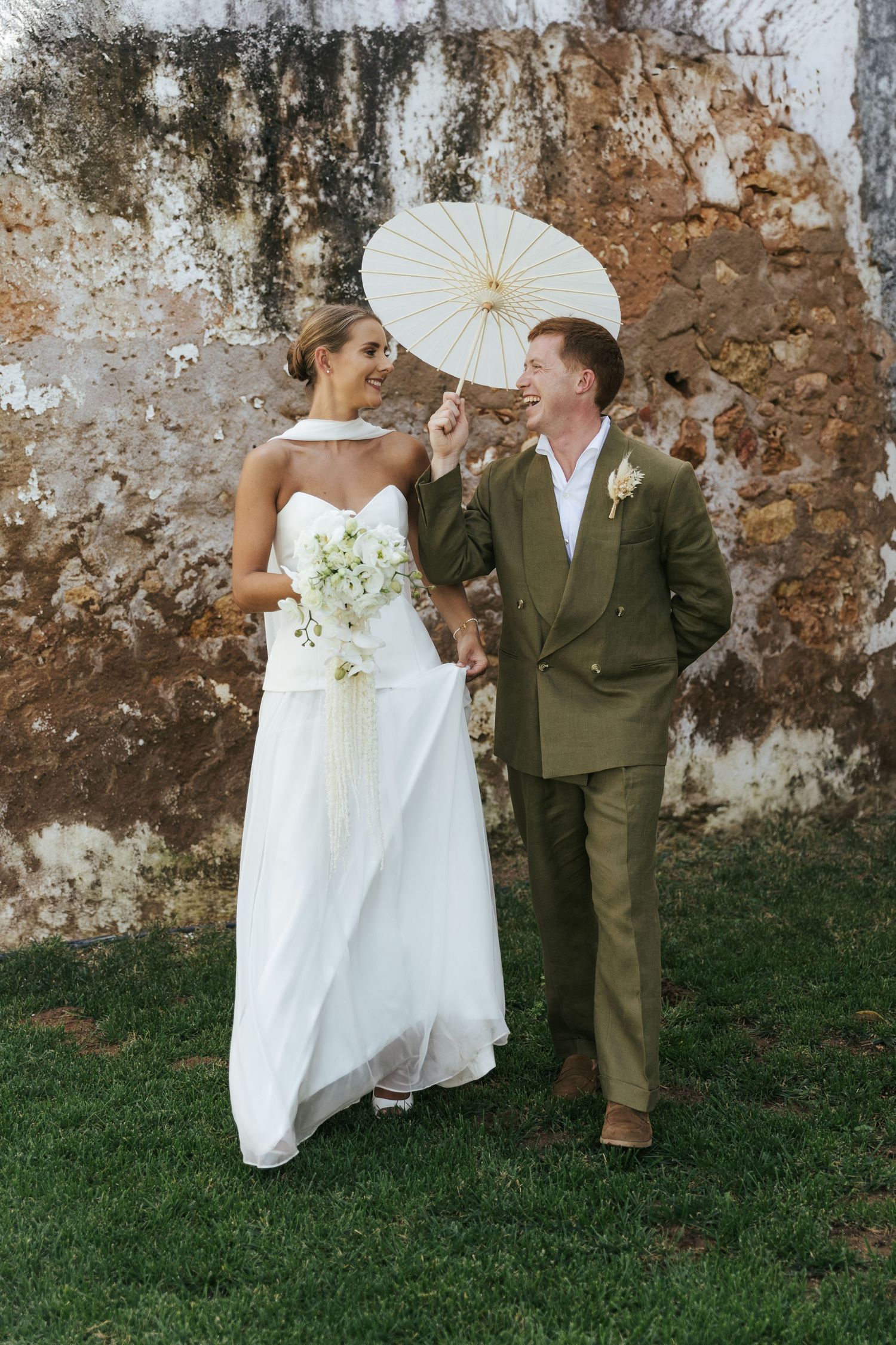 Bride in white gown and groom in olive suit hold parasol and bouquet, laughing together against rustic stone wall.