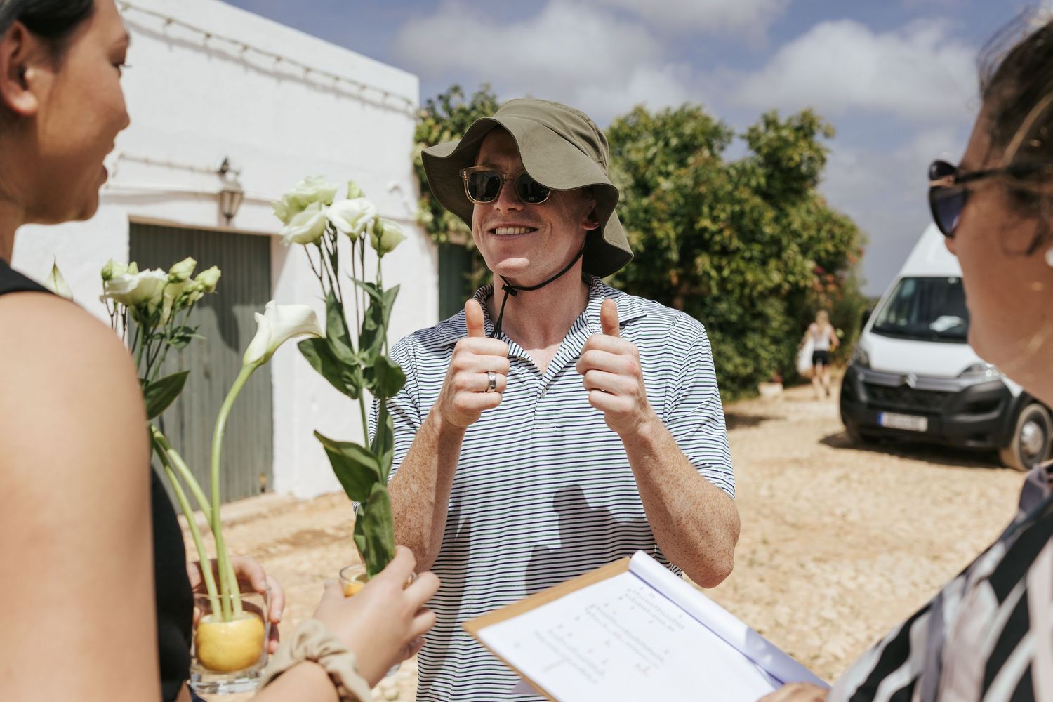 Man giving thumbs up while holding clipboard outdoors, talking with women, one holding white flowers, on a sunny day.