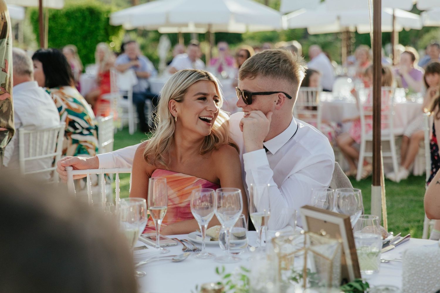 Couple shares a candid moment at an elegant outdoor wedding reception under white umbrellas.
