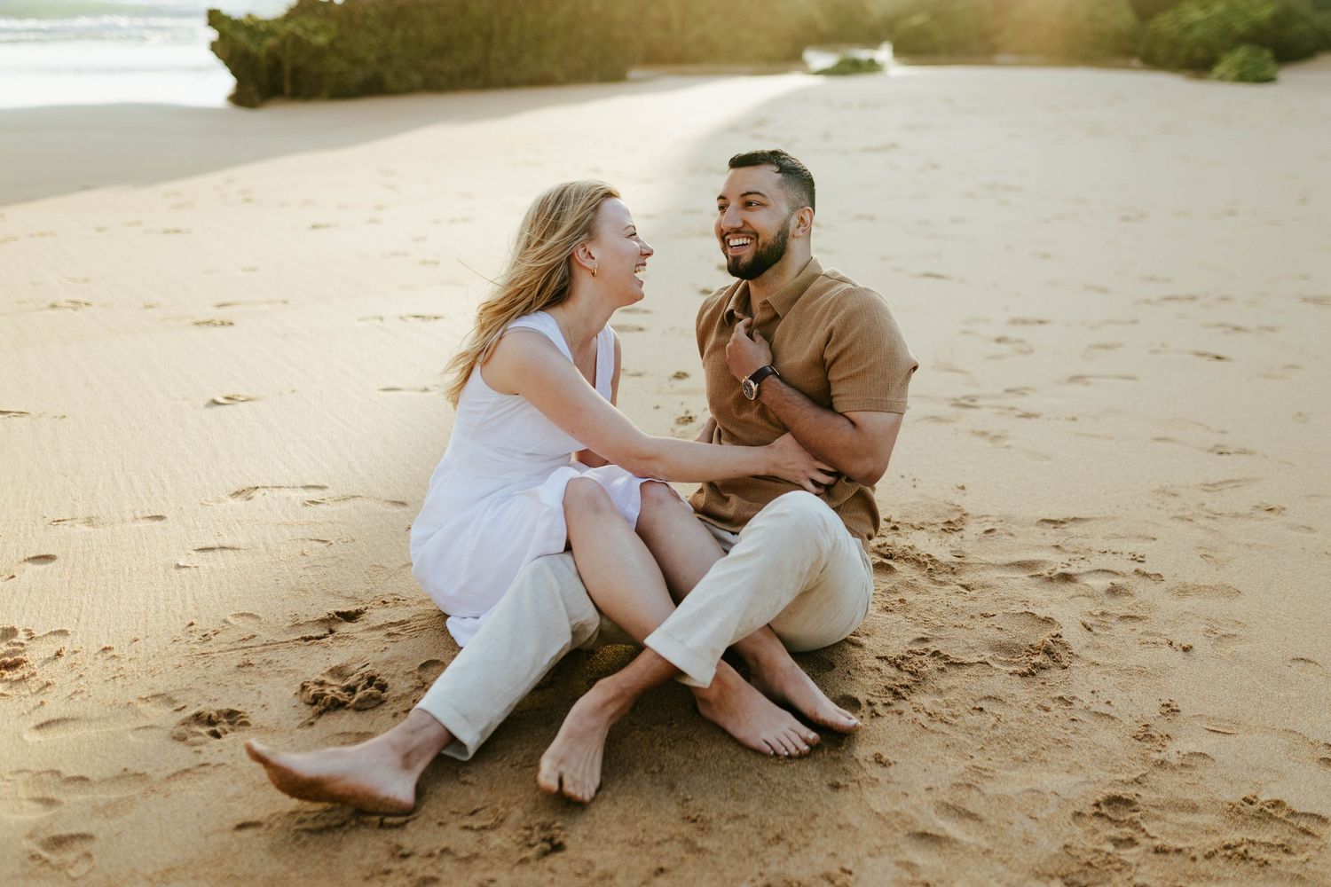 A laughing couple sits together on a sandy beach during a golden afternoon.