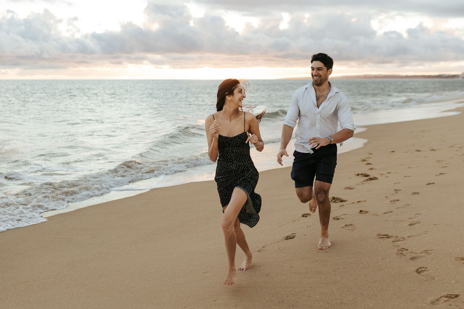 A couple runs joyfully together along a sandy beach at sunset with waves crashing beside them.