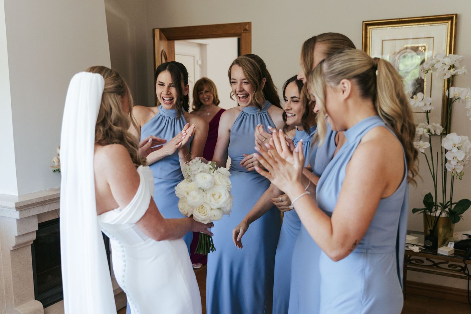Bride in white gown holds bouquet as bridesmaids in blue dresses clap and smile during wedding preparations.