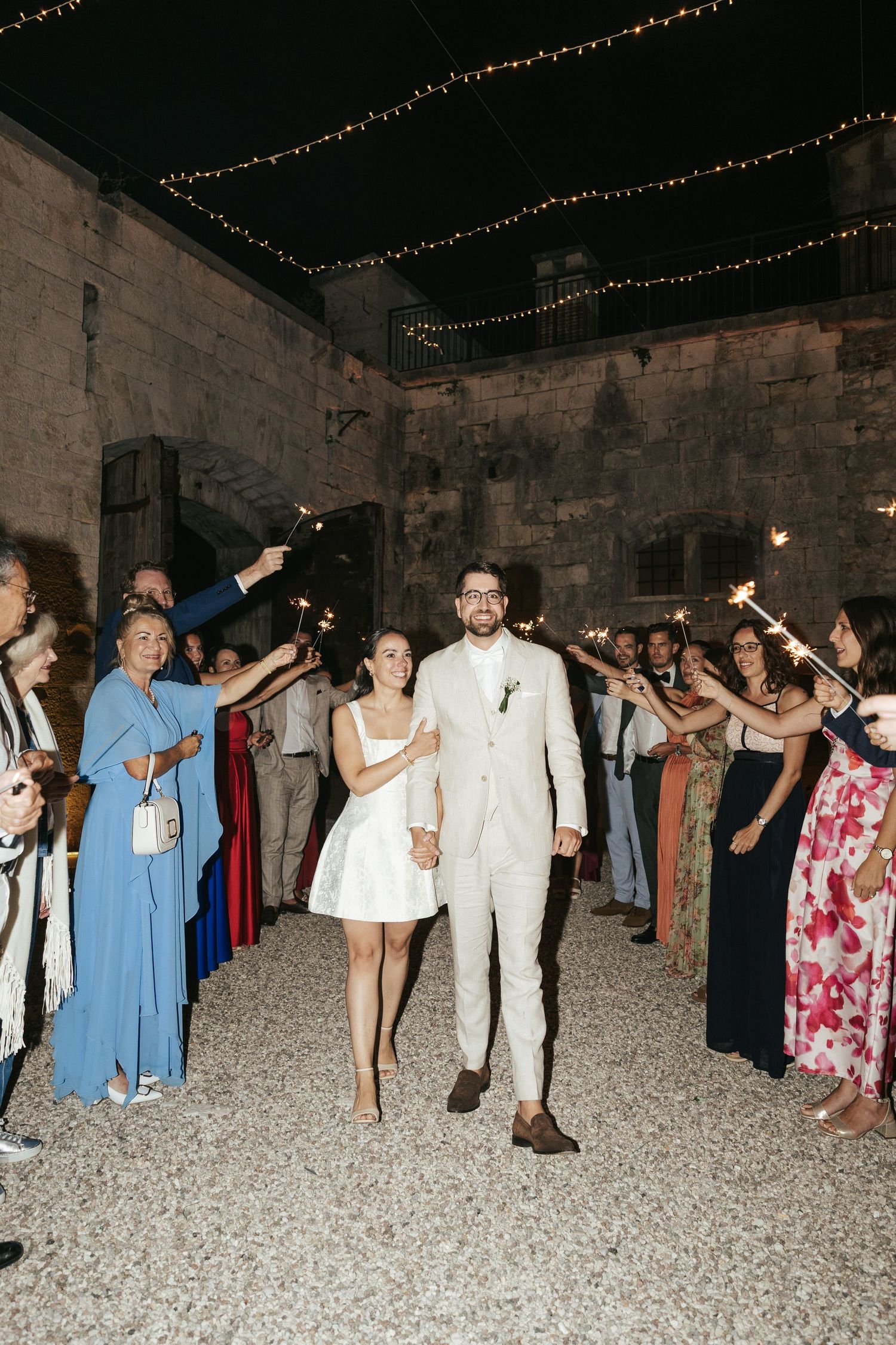 Wedding guests dance in a line on a stone patio with string lights overhead during an evening reception celebration.