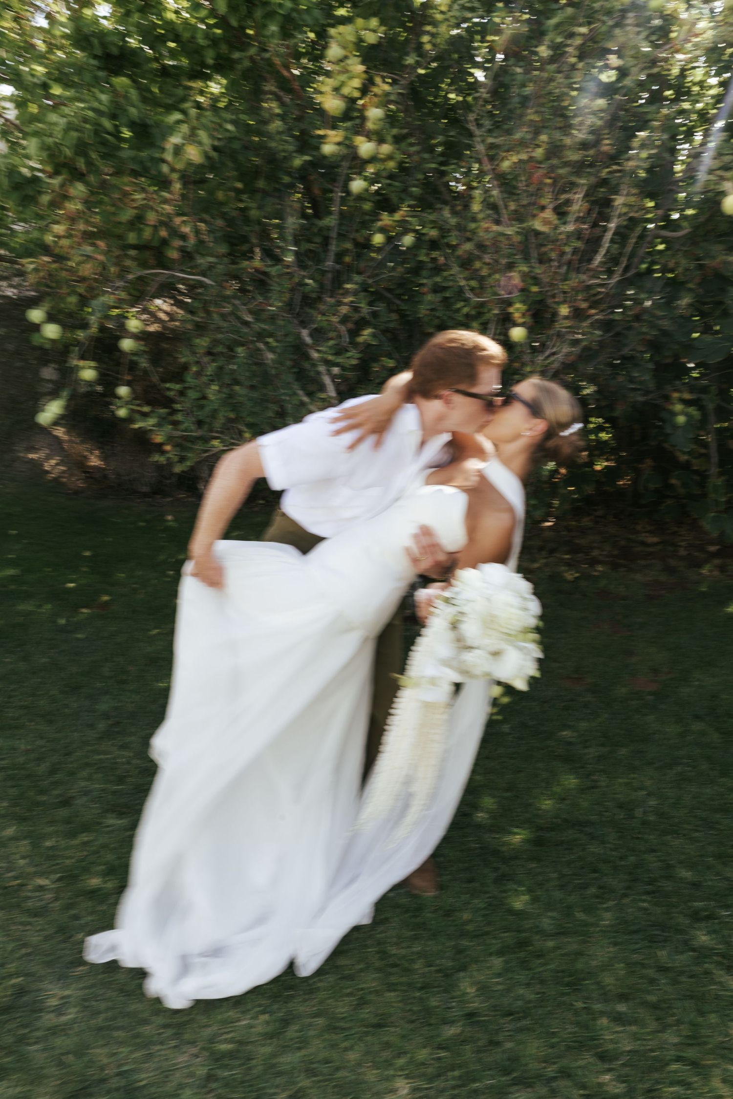 A romantic wedding couple dips in a graceful dance pose on a green lawn surrounded by trees and foliage.