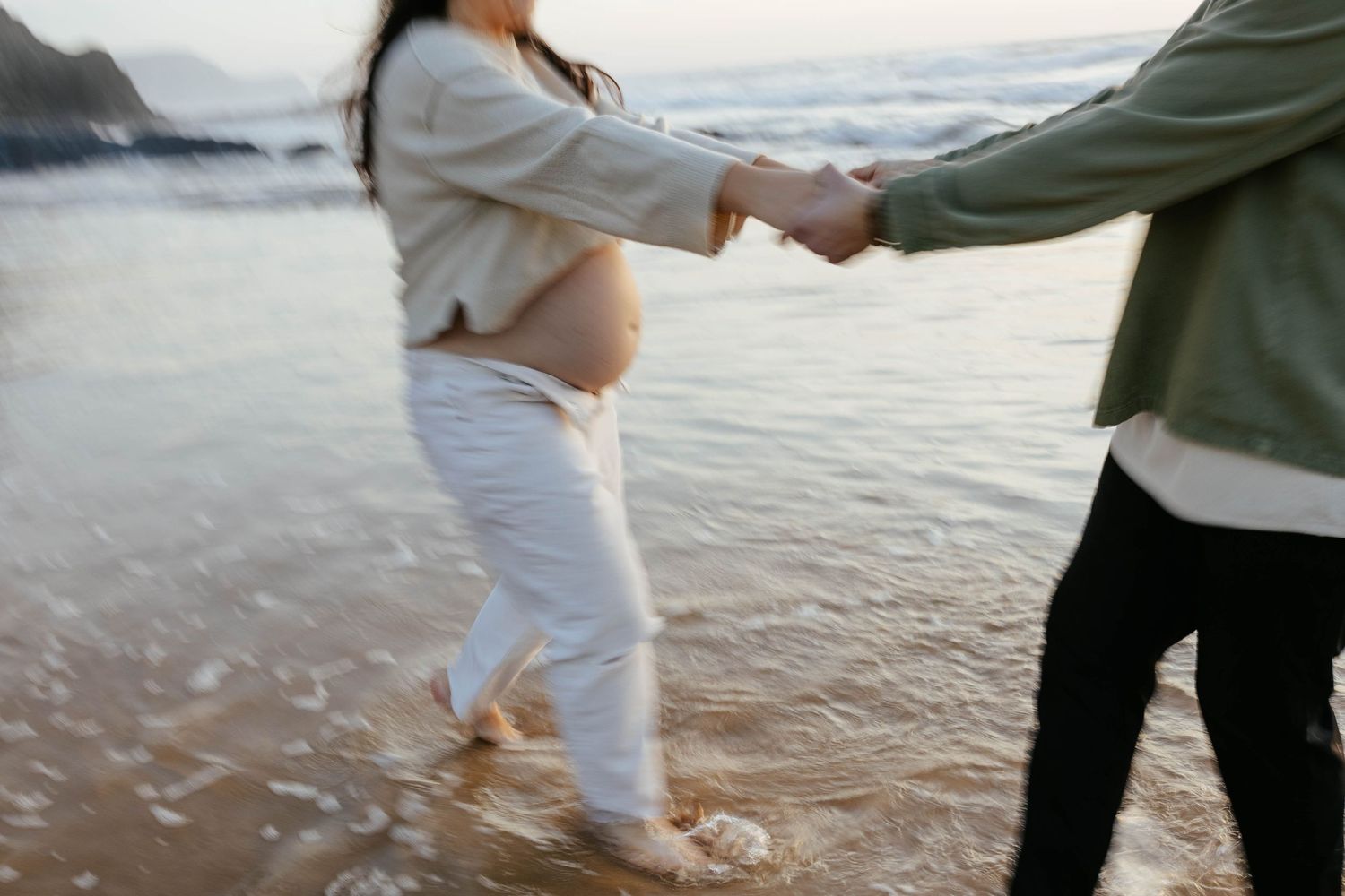 A couple holds hands while walking along the shoreline at sunset with misty mountains in the background.