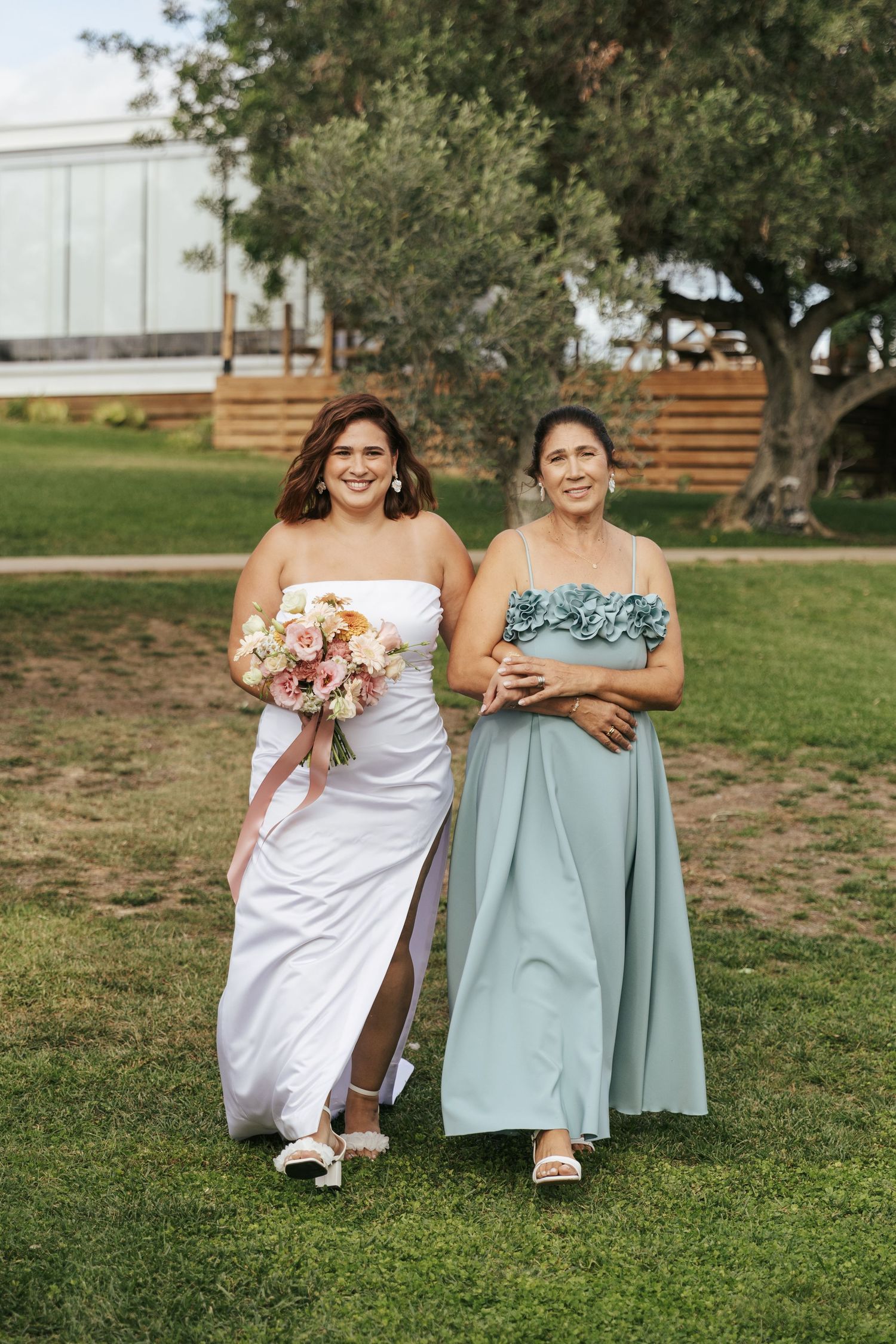 Two people walk together on grass, one wearing a white wedding gown and holding a bouquet.
