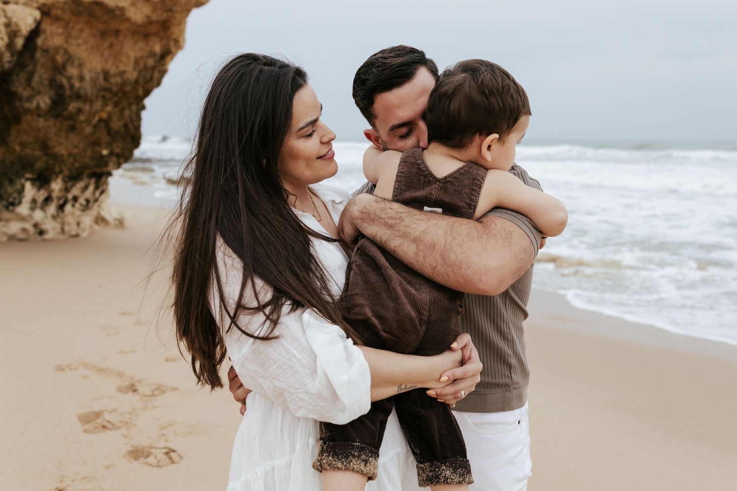 Family sharing a loving embrace on a scenic beach with rocky cliffs.