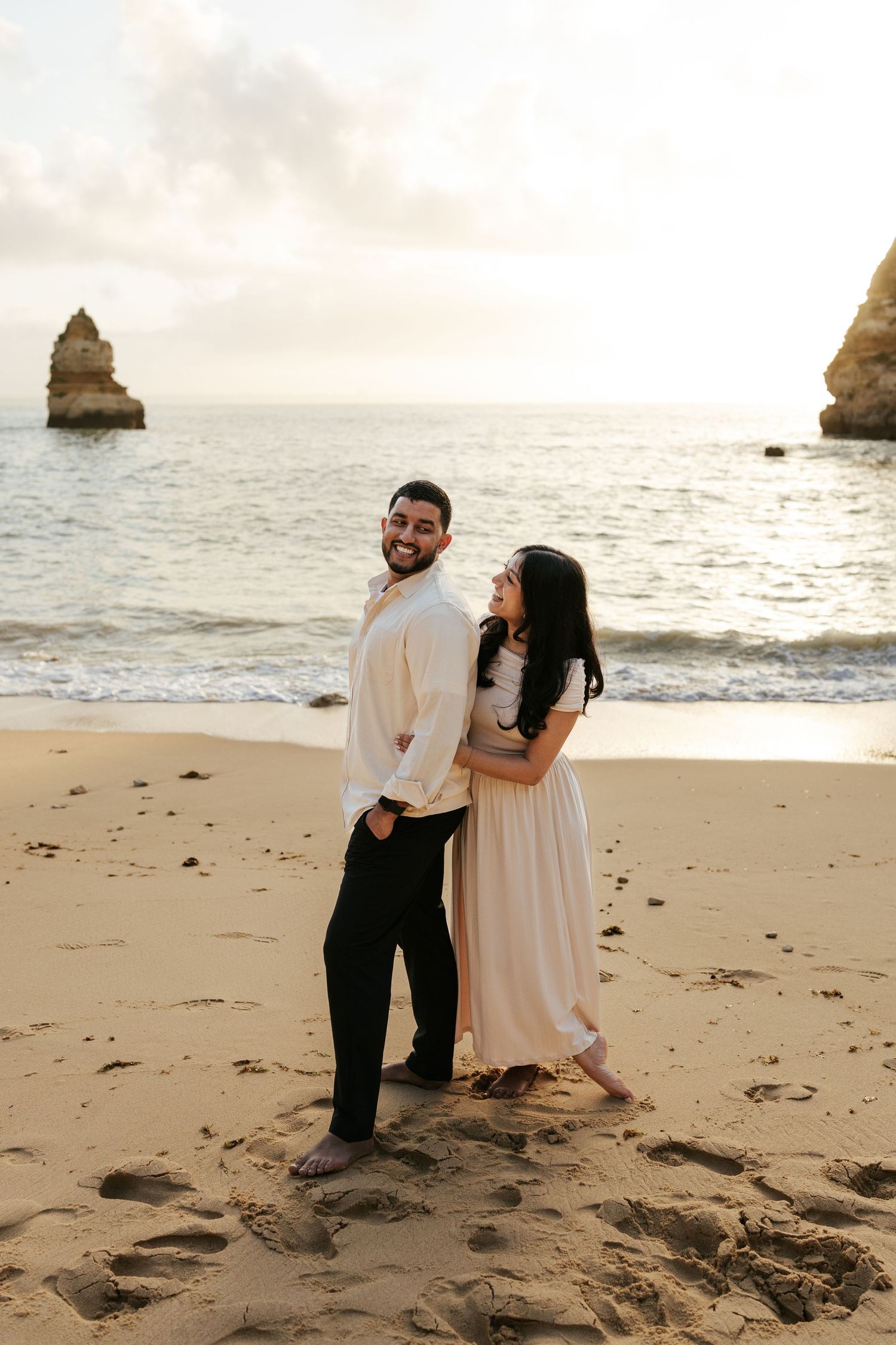 Couple stands together on a sandy beach at sunset with dramatic rock formations in the ocean background.