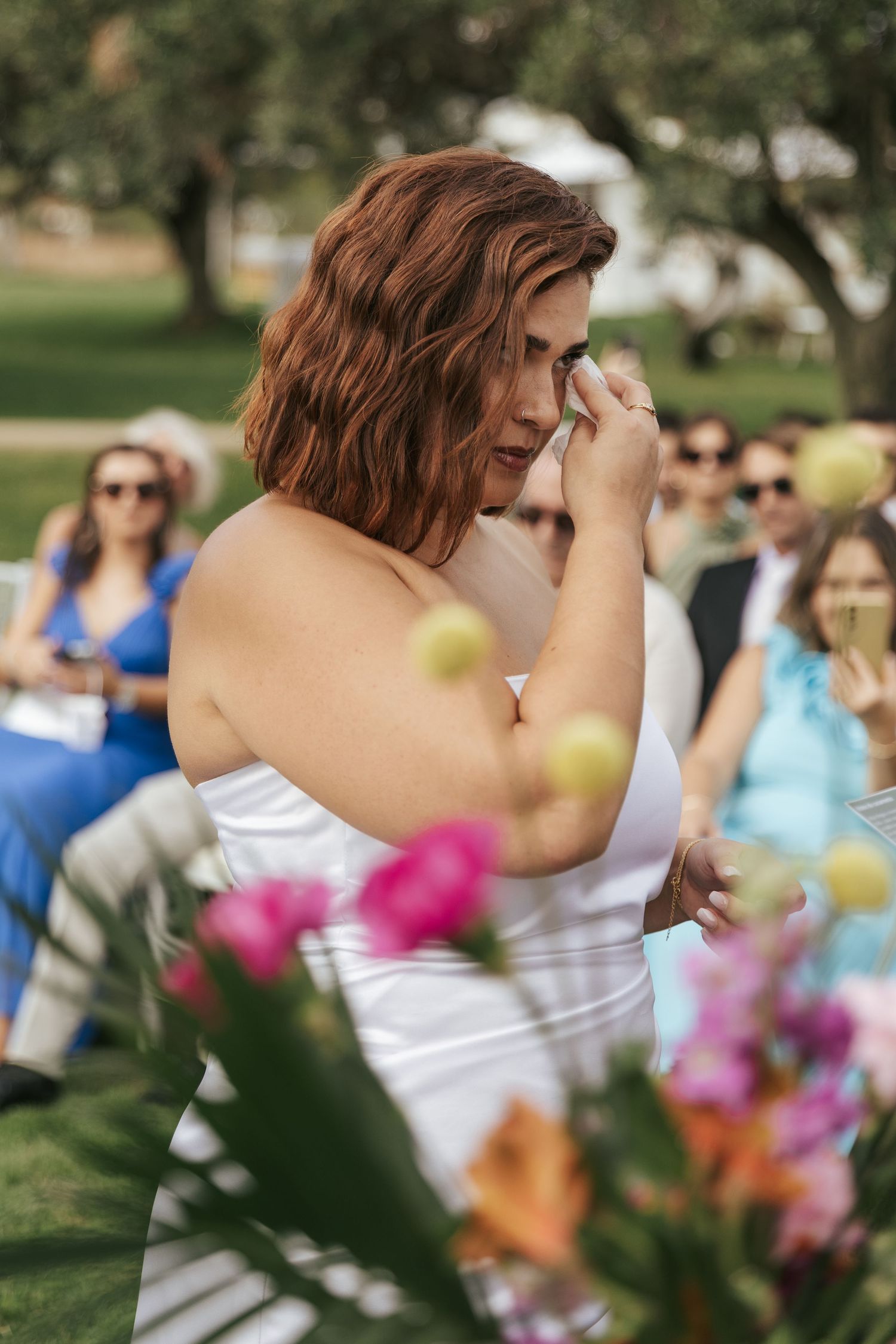A person in a white dress wipes away tears during an emotional outdoor celebration with colorful flowers in the foreground.