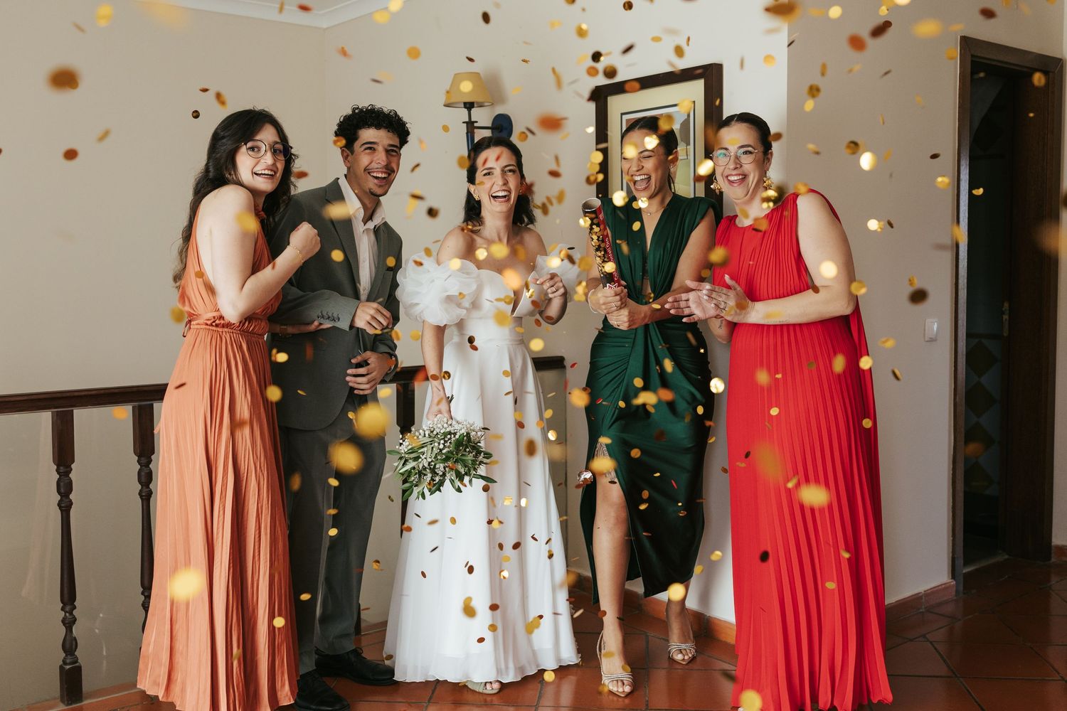 A group of bridesmaids in coral and red dresses pose together at a wedding celebration with confetti falling.