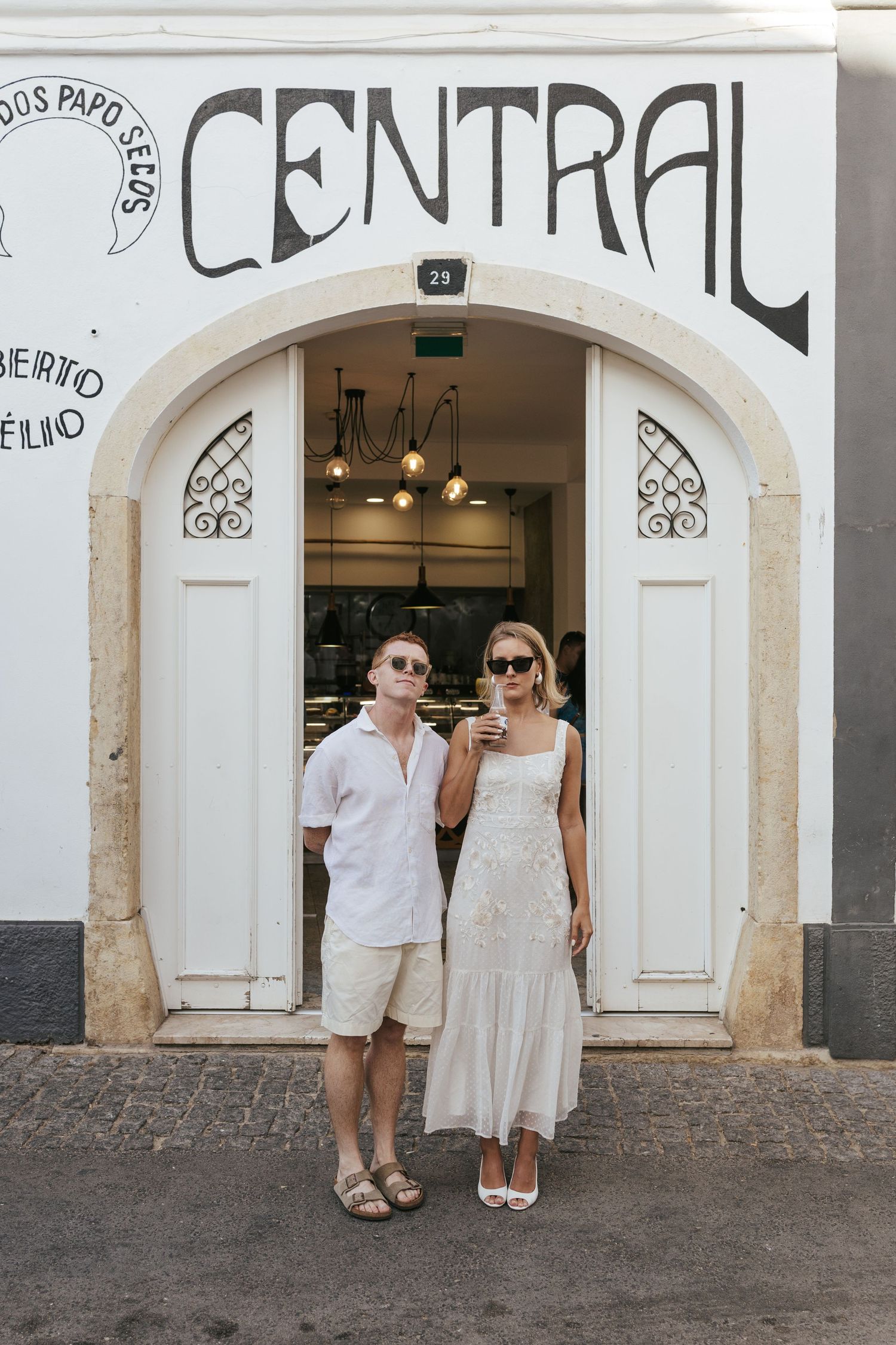 A couple stands in front of a white arched storefront with 'Padaria Central'. Lagos, Portugal.