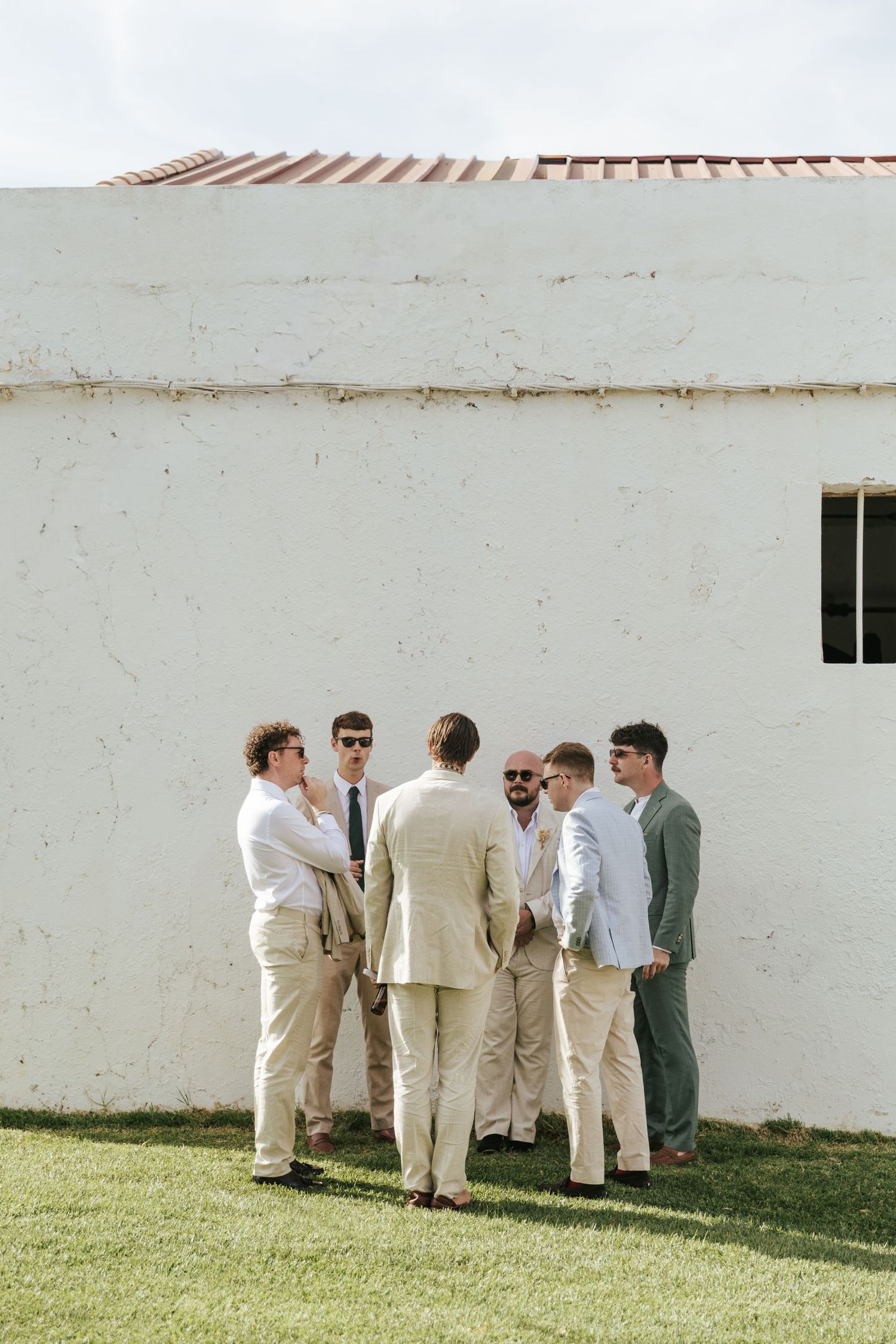 Group of groomsmen in neutral suits gathered outside a white stucco building on a sunny wedding day.