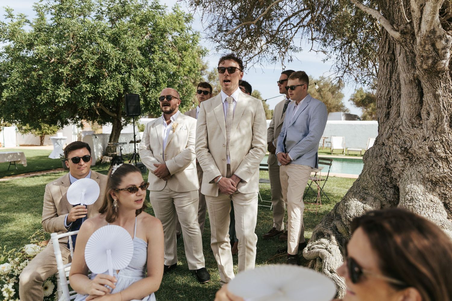 Outdoor wedding ceremony with groom and groomsmen standing under a tree while guests are seated in white chairs on grass.