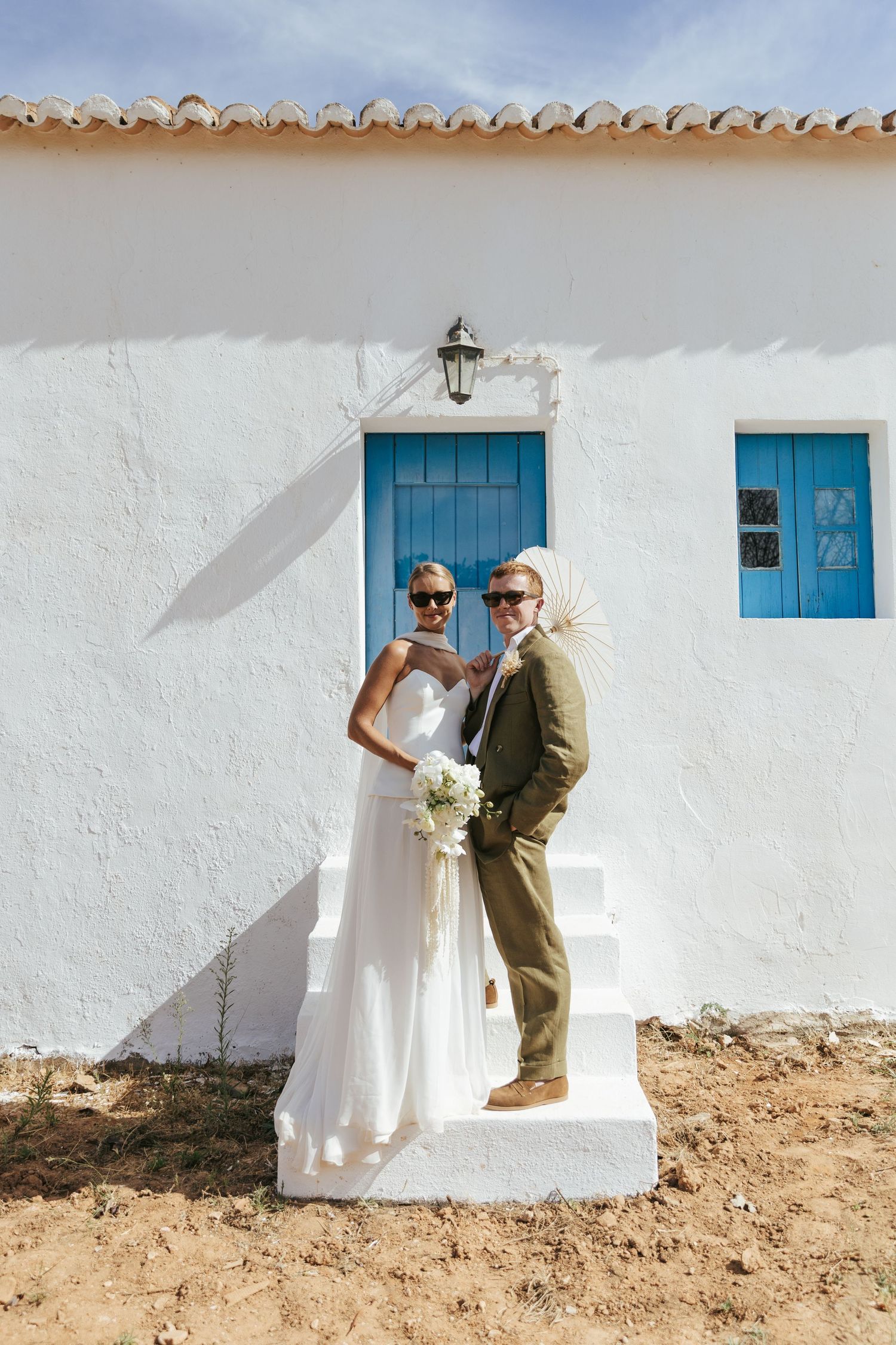 Bride and groom in sunglasses pose on white steps of Mediterranean-style building with blue doors and sandy ground.
