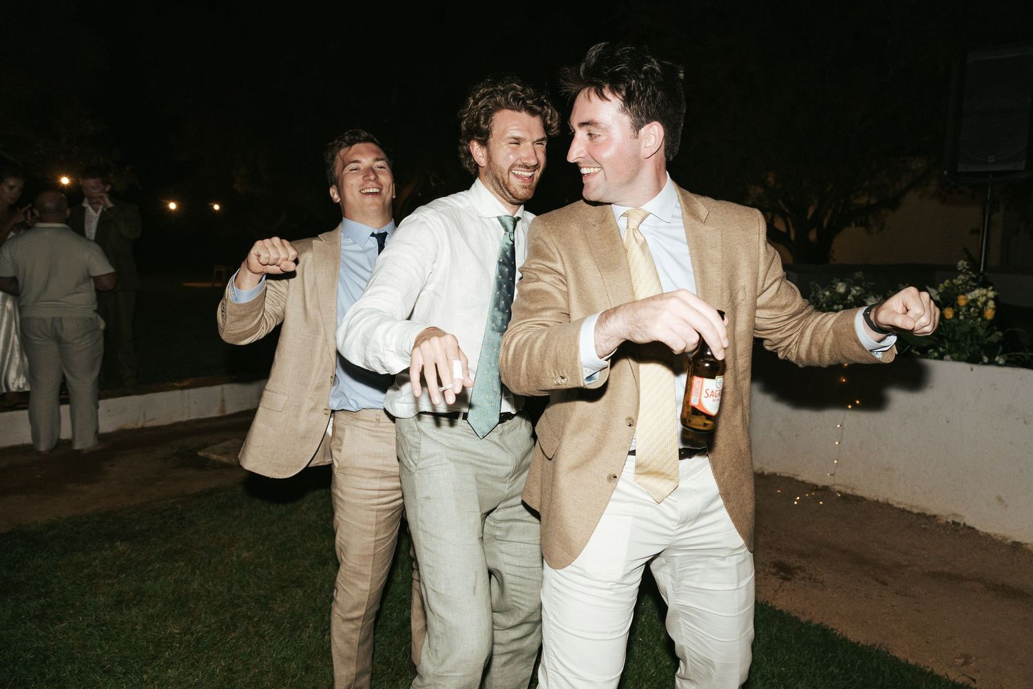 Three men in tan blazers and ties laugh and dance outdoors at a nighttime celebration, holding drinks.