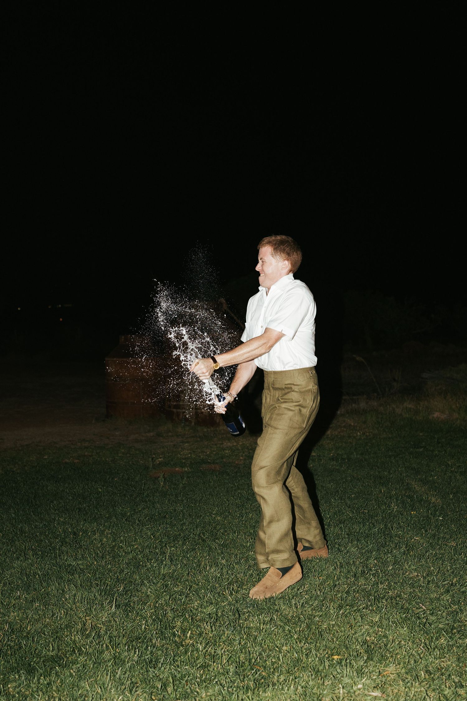 Man spraying champagne bottle on dark lawn during outdoor wedding celebration at night.