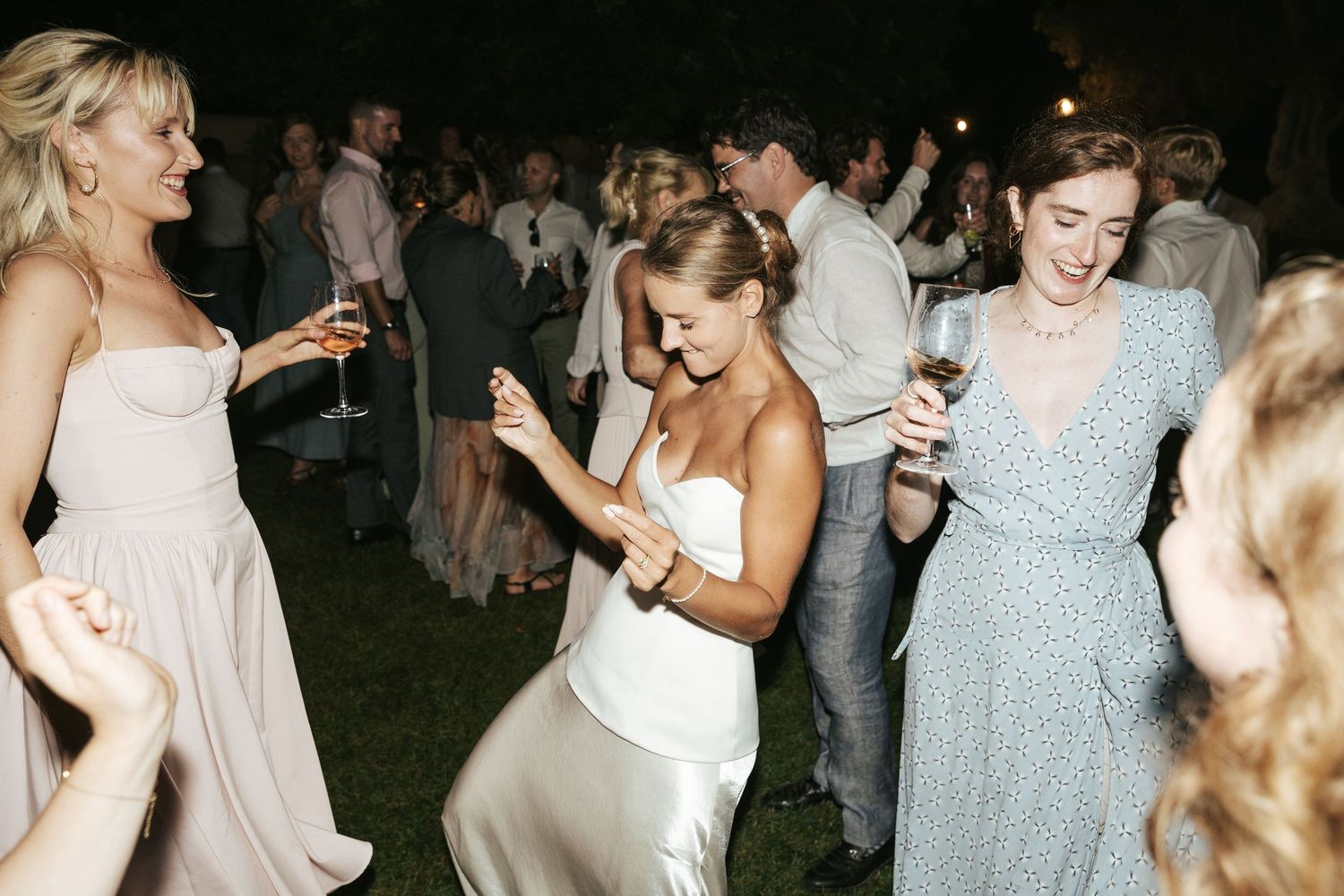 Bride in white satin dress dancing joyfully with guests at outdoor evening wedding reception.