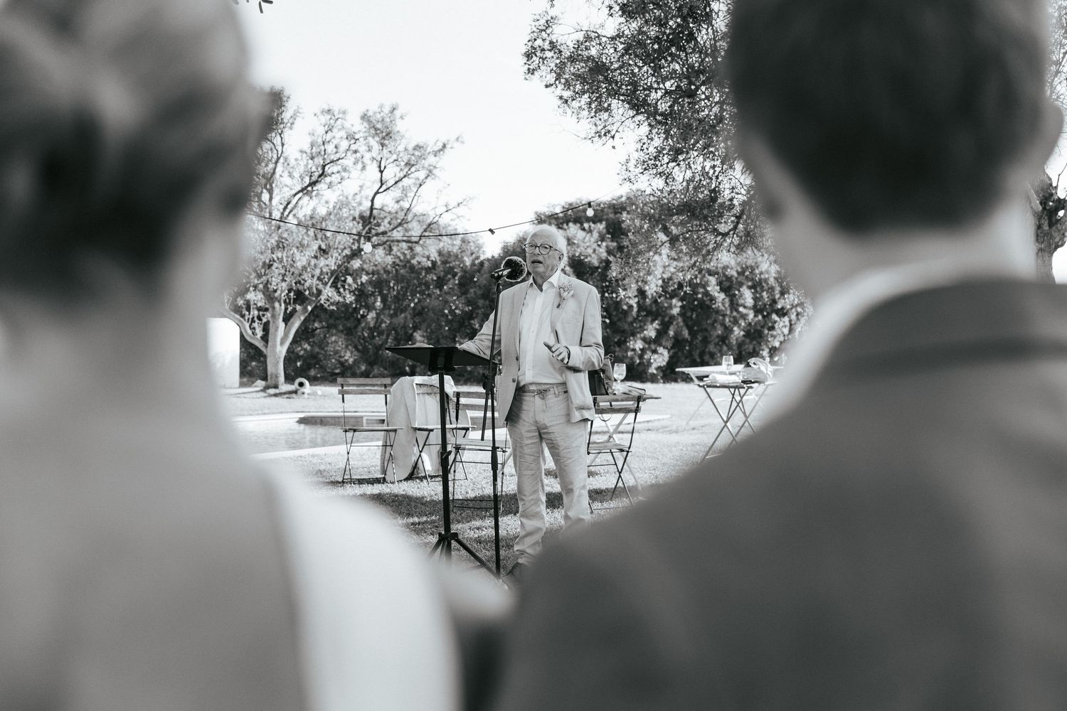 Black and white photo of a man speaking at an outdoor wedding ceremony before seated guests.