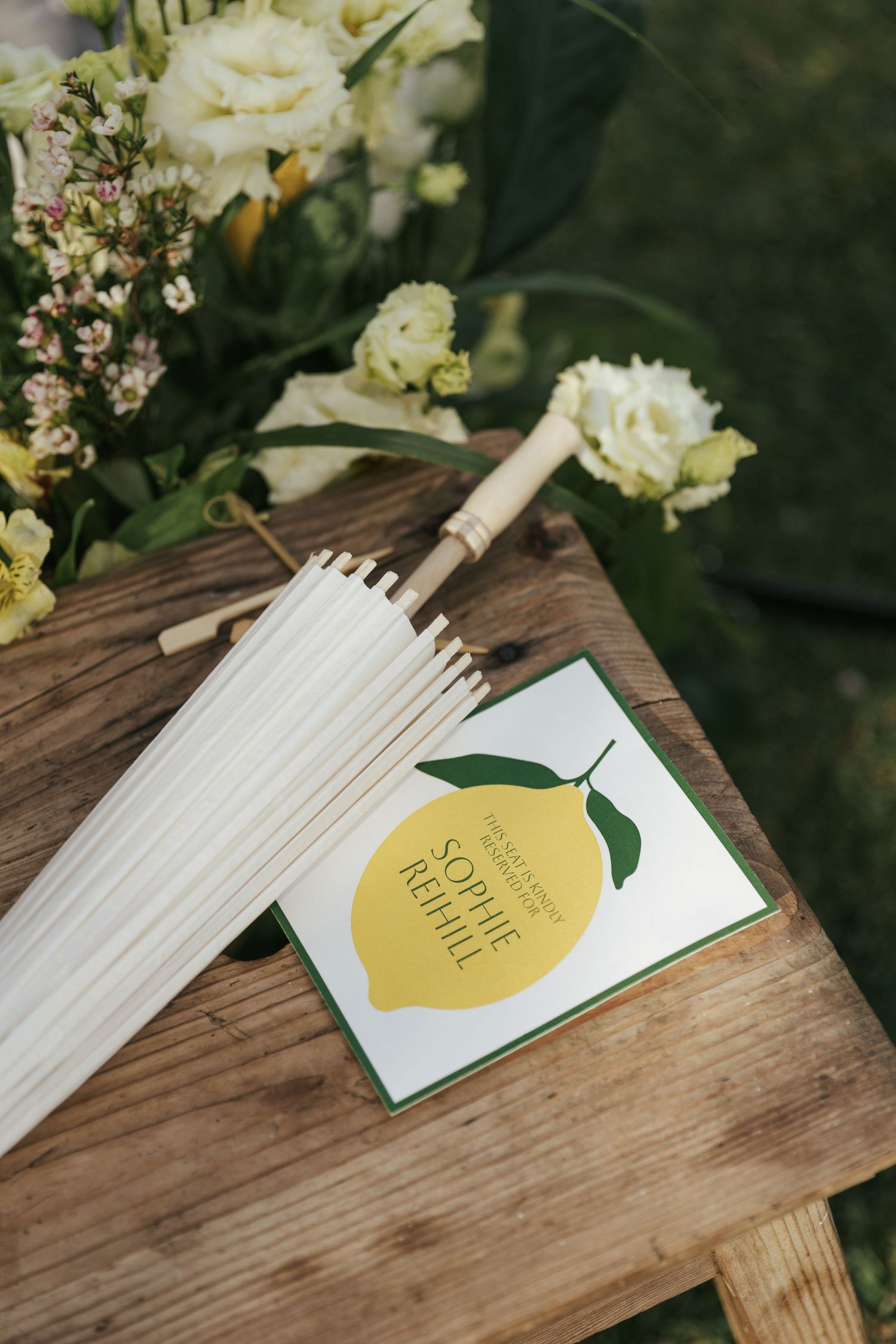 Wedding program with lemon design on wooden bench beside white parasol and fresh floral arrangement.
