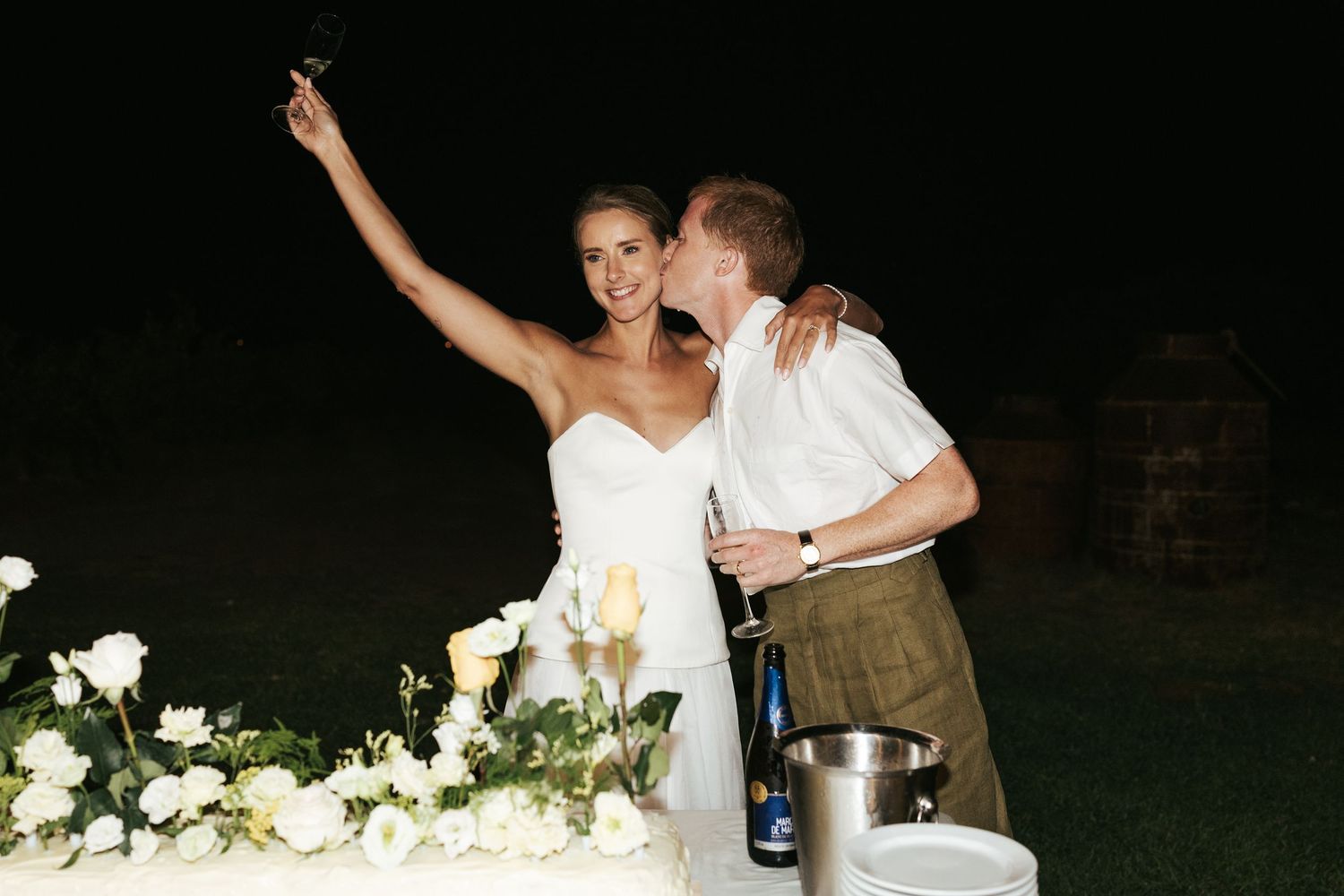 Bride raising wine glass beside groom near large floral wedding cake at night.