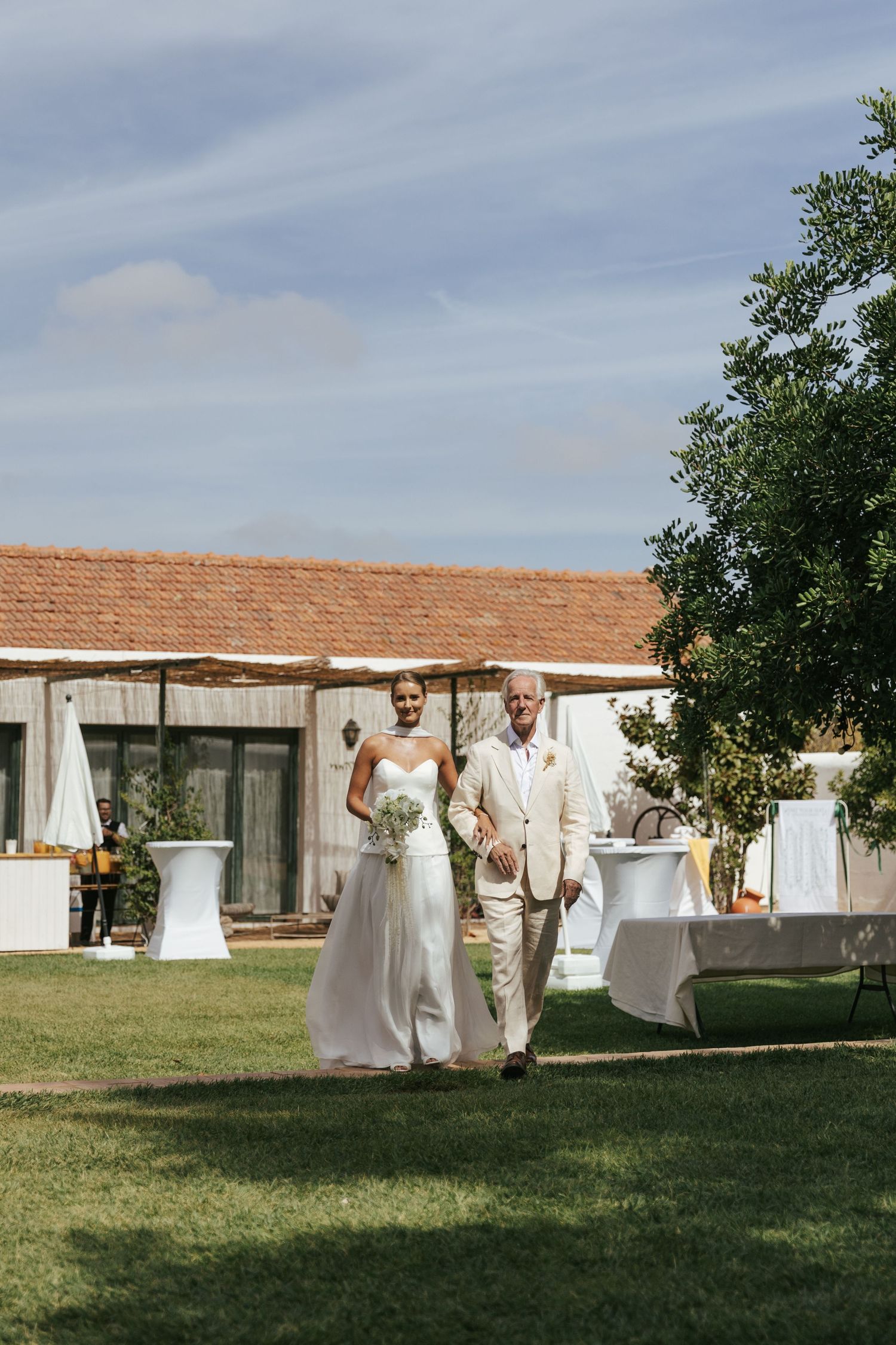 Bride in white gown walks with older man in tan suit across green lawn at outdoor wedding venue.