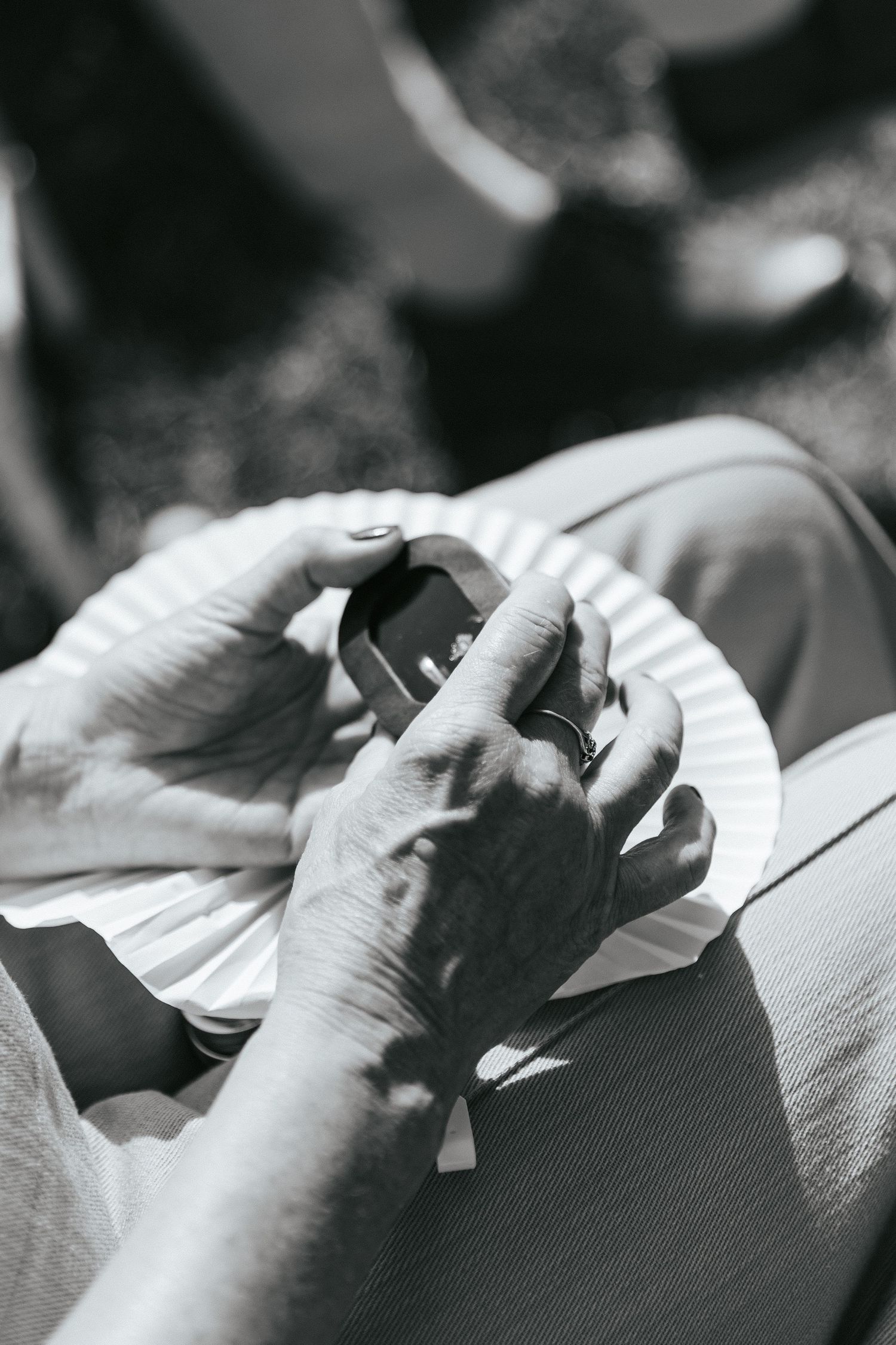 Black and white close-up of elderly hands holding smartphone and paper plate outdoors.