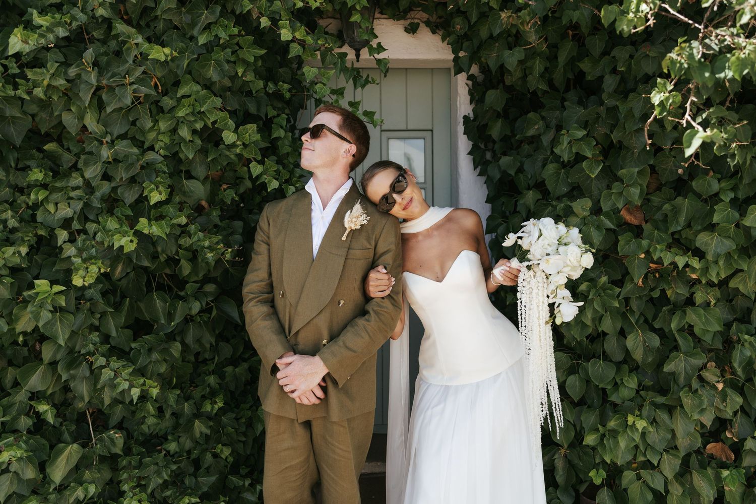 Bride in white strapless gown with bouquet leans on groom in olive suit, both wearing sunglasses before ivy-covered wall.