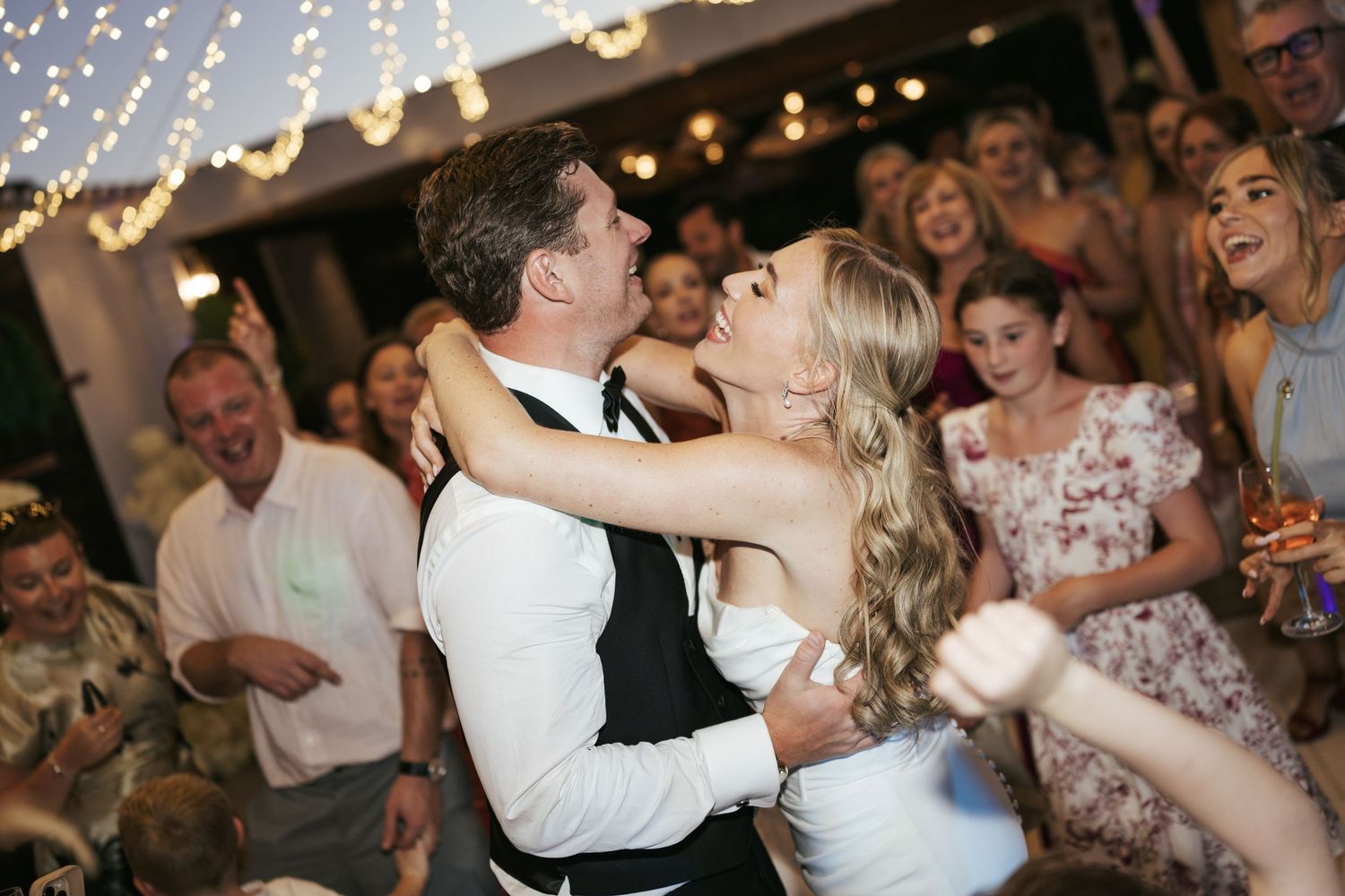 Bride and groom share romantic first dance at wedding reception with guests celebrating in background under fairy lights.