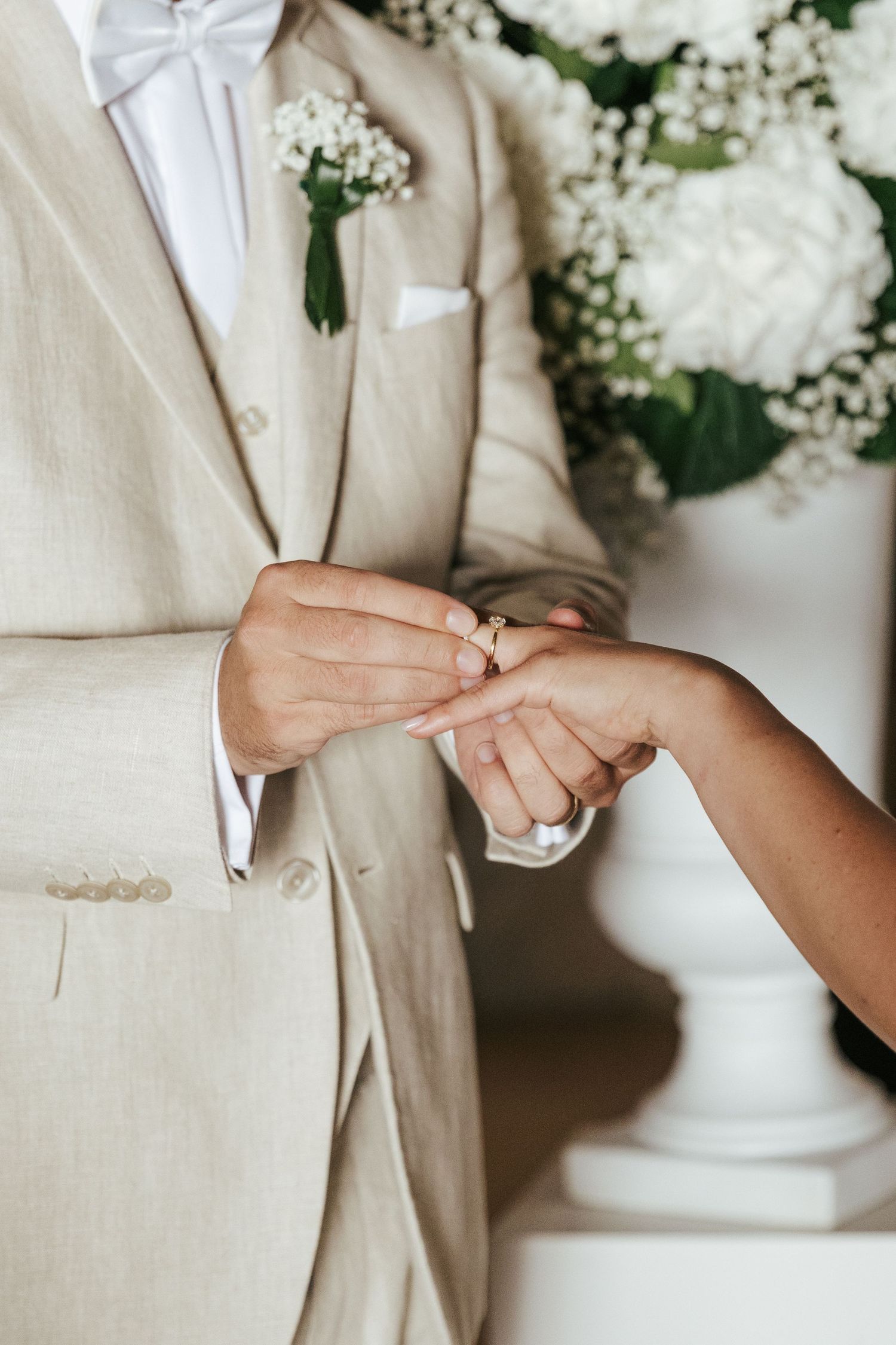 Close-up of hands exchanging wedding rings with white floral arrangement in background.