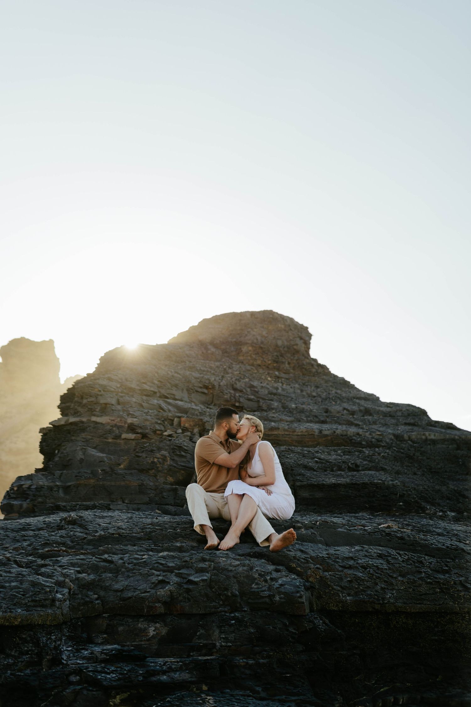 Two figures embrace while sitting on rocky coastal cliffs at sunset, creating a romantic silhouette against the sky.
