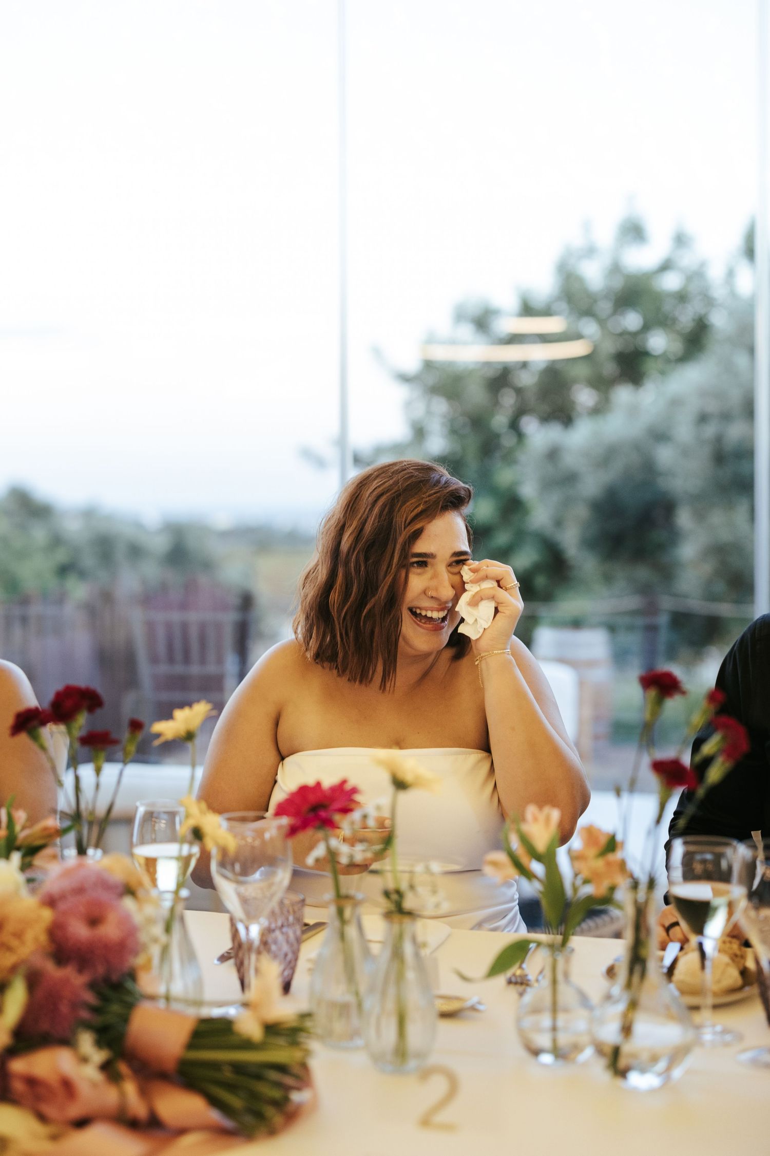 Someone in a white dress smiles while sitting at an elegant table decorated with pink and red flowers for a celebration.