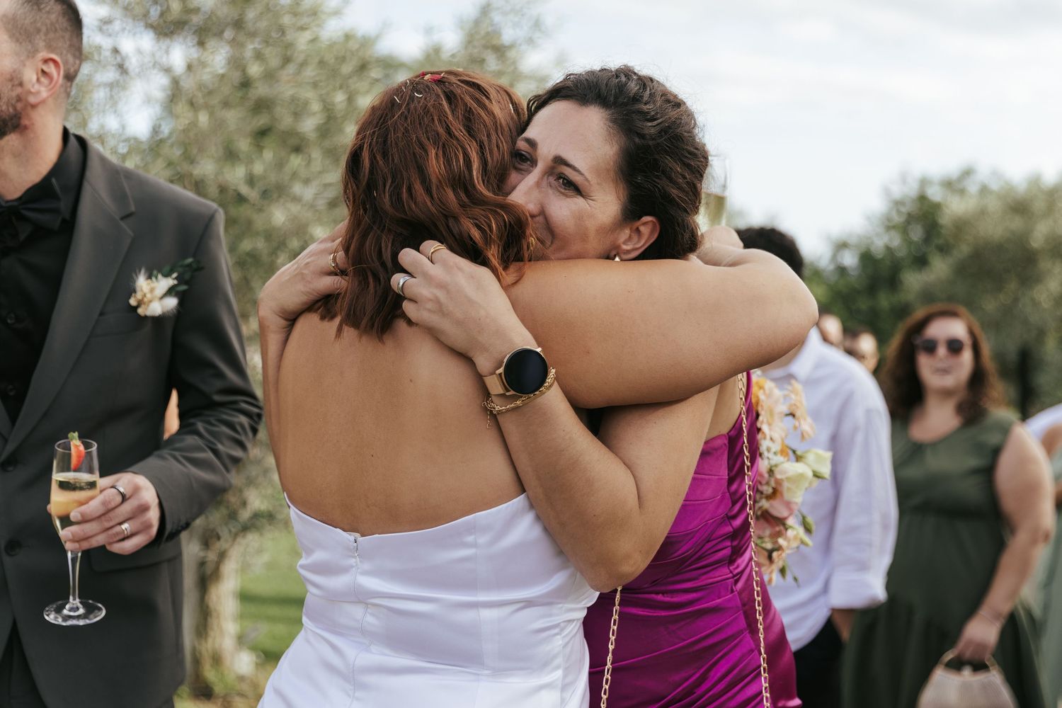 Two guests share an emotional embrace at an outdoor wedding celebration while others mingle in the background.