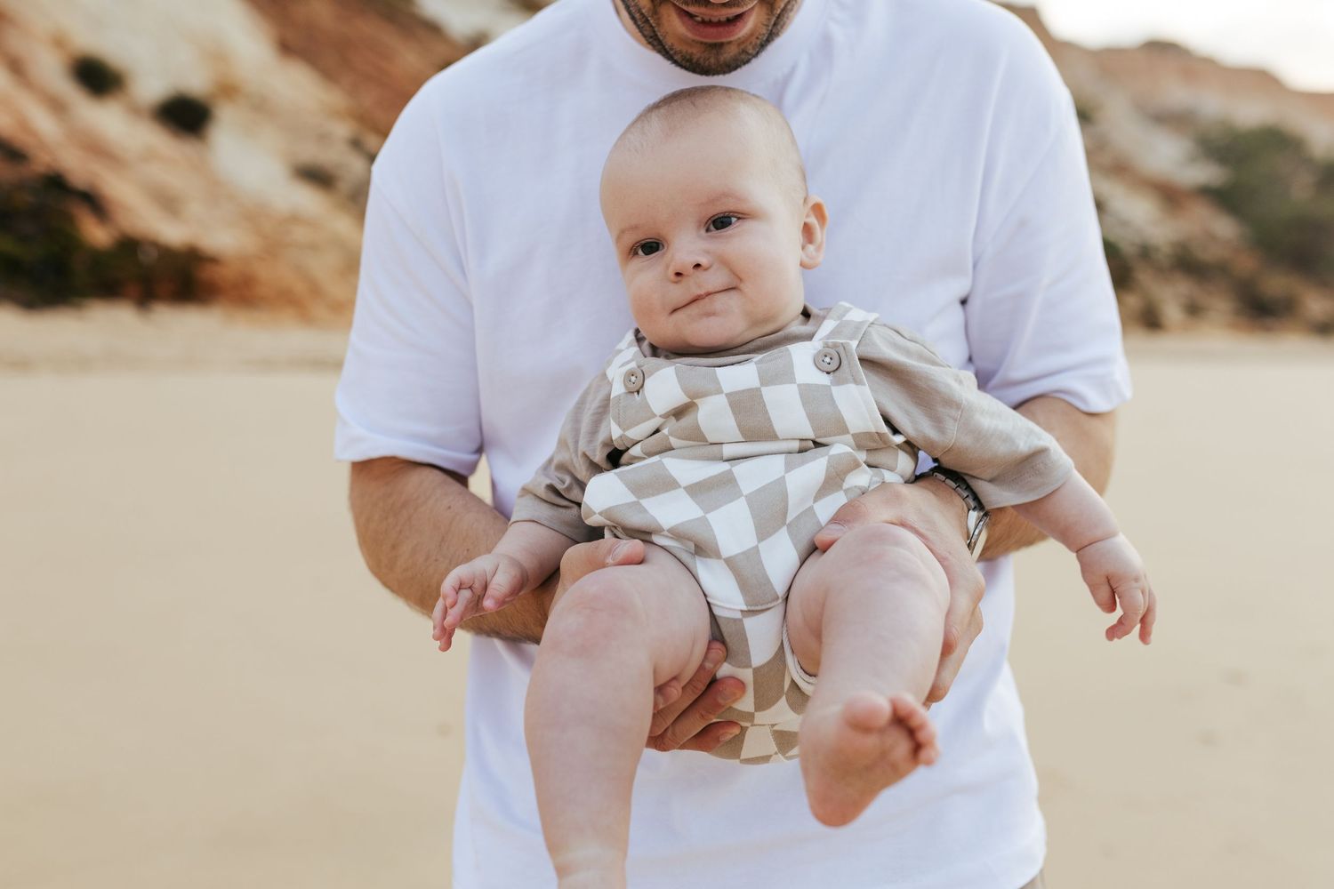A baby wearing a striped romper sits supported in someone's arms against a desert landscape background.