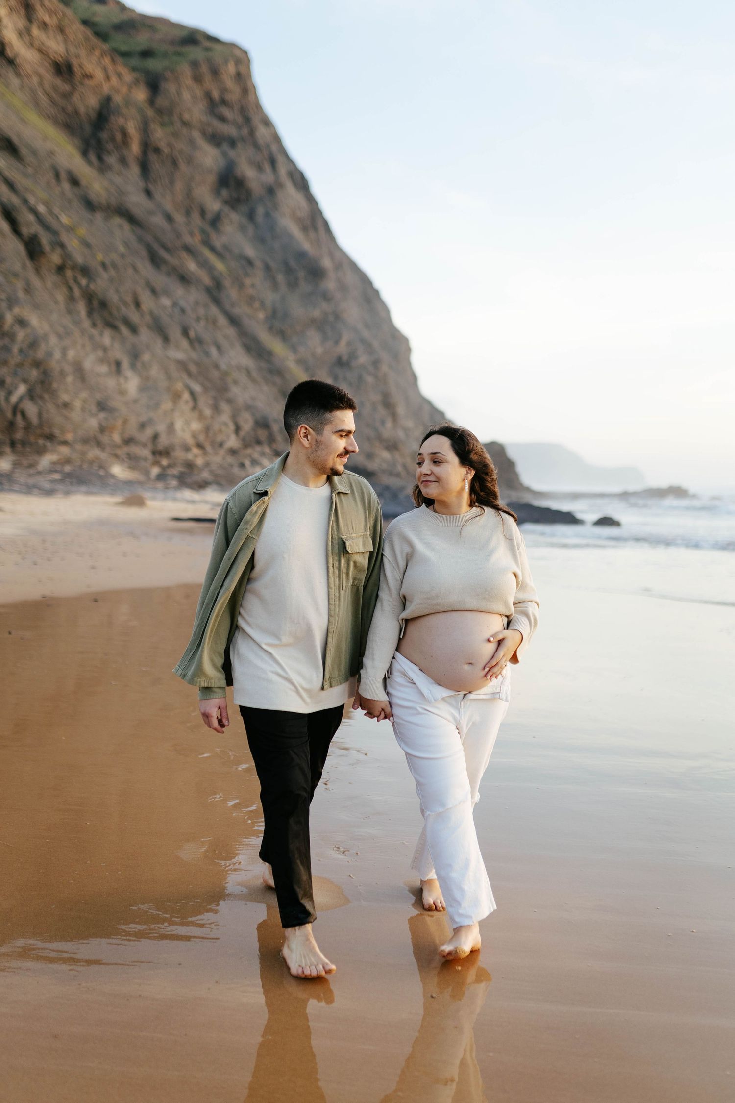 Couple taking romantic barefoot stroll along sandy beach with towering coastal cliffs at golden hour.