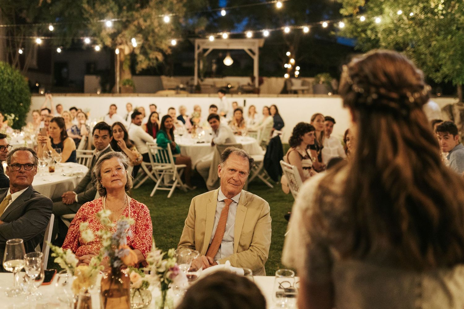 Wedding guests seated at outdoor reception tables listen attentively during evening speeches under string lights.