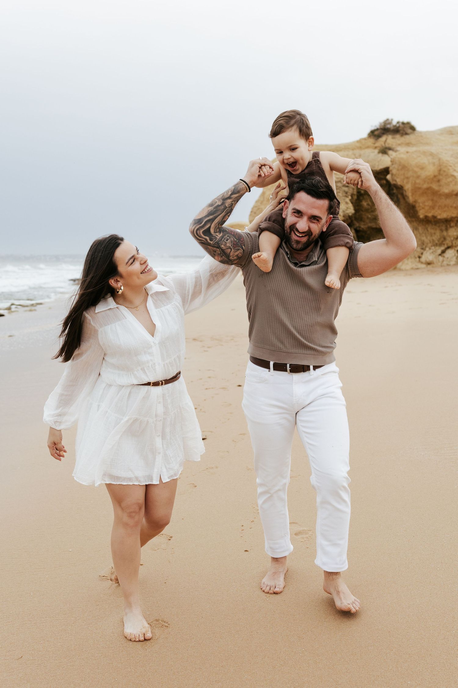 A family walks barefoot along a sandy beach together on a cloudy day by rocky cliffs.