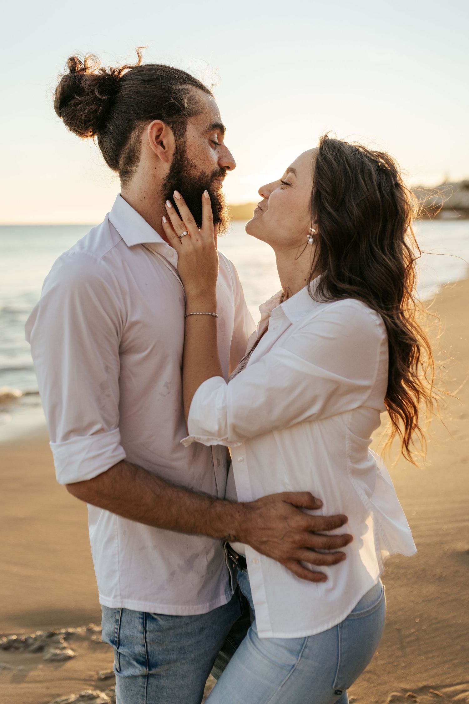 A romantic couple embraces on a beach at sunset wearing white shirts and casual attire.