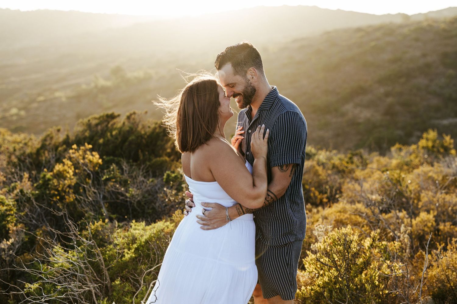 A couple embraces on a hilltop at golden hour surrounded by wildflowers and mountain scenery.