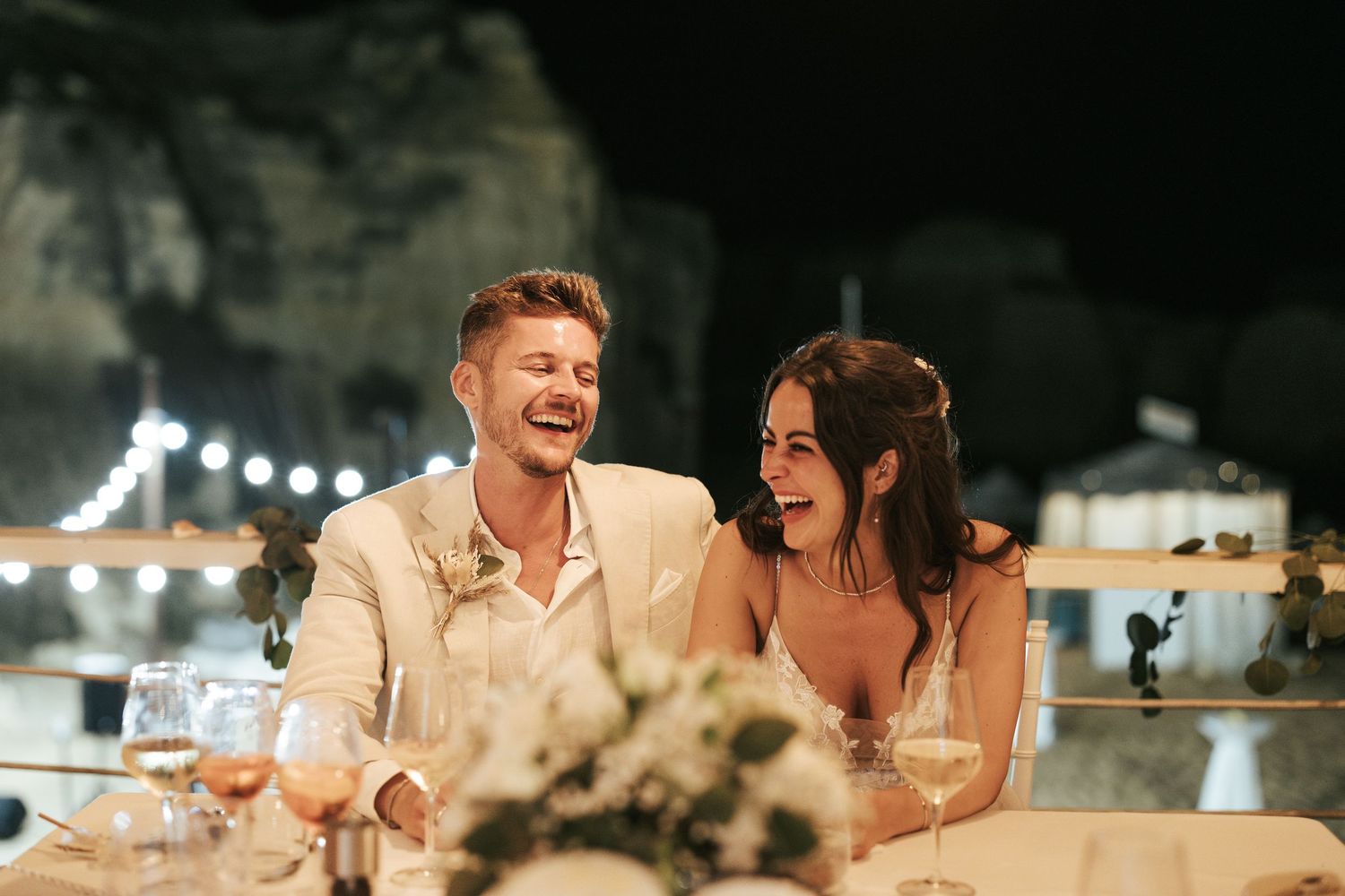 Couple sharing a candid laughing moment while dining at an outdoor evening venue with string lights in background.