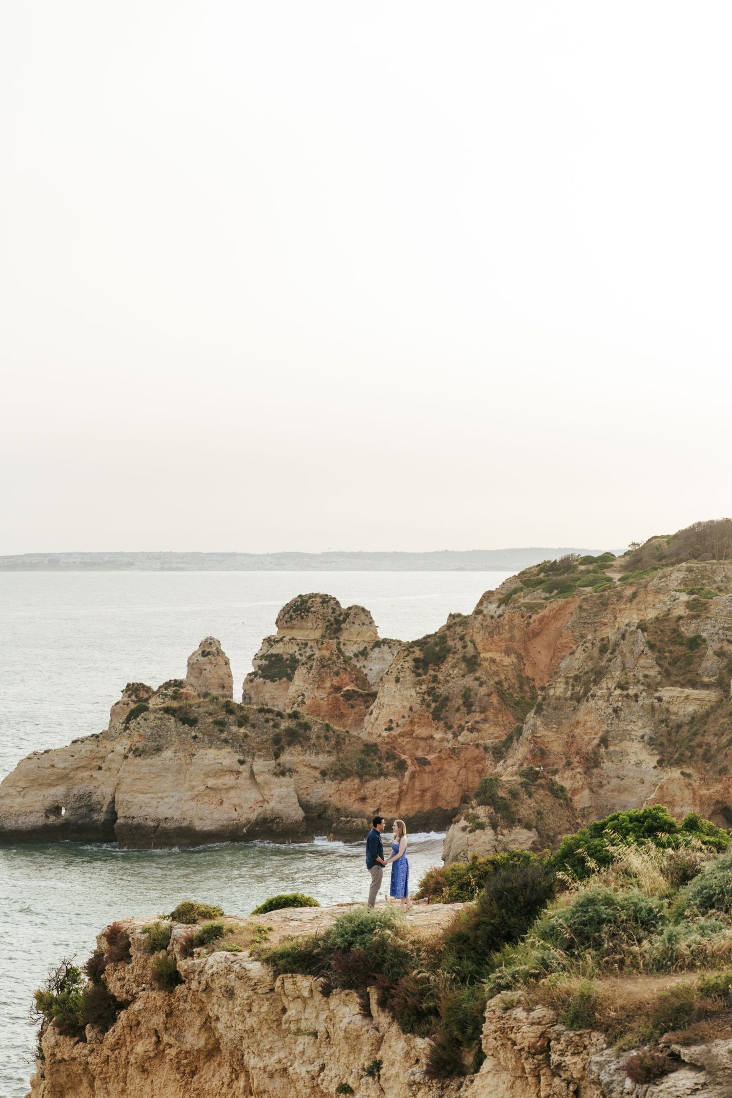 Couple standing on rocky coastal cliffs overlooking the ocean, surrounded by rugged sandstone formations and green shrubs.