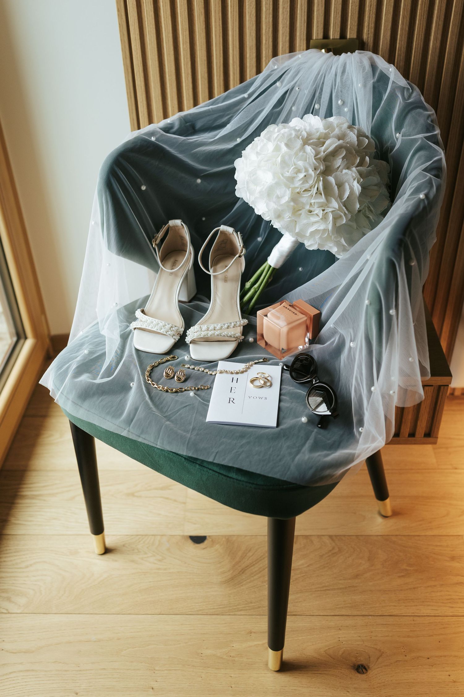 Wedding accessories including shoes and veil arranged on a gray velvet chair.