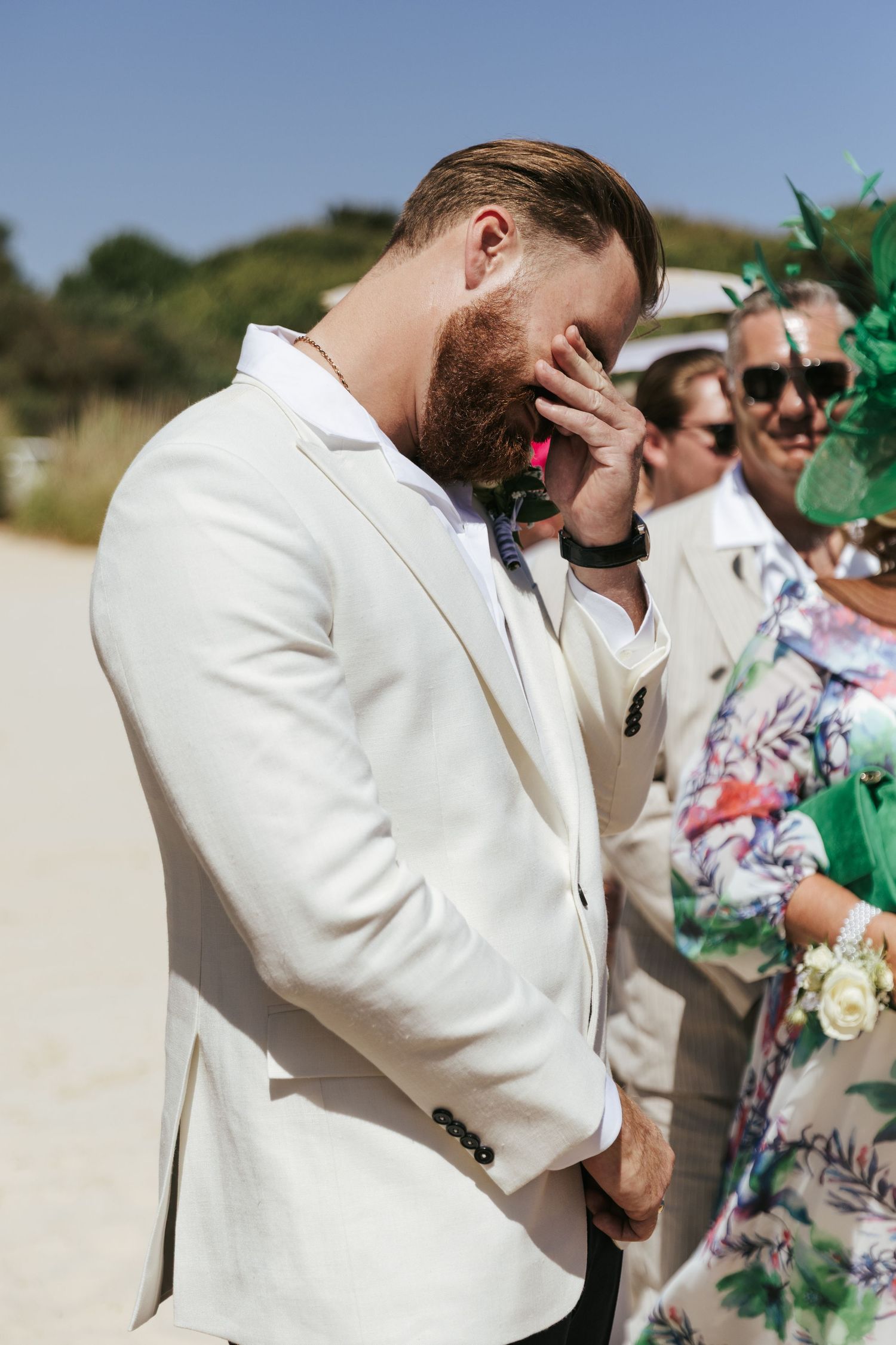A person in a white suit jacket wipes tears away during an emotional outdoor wedding ceremony.
