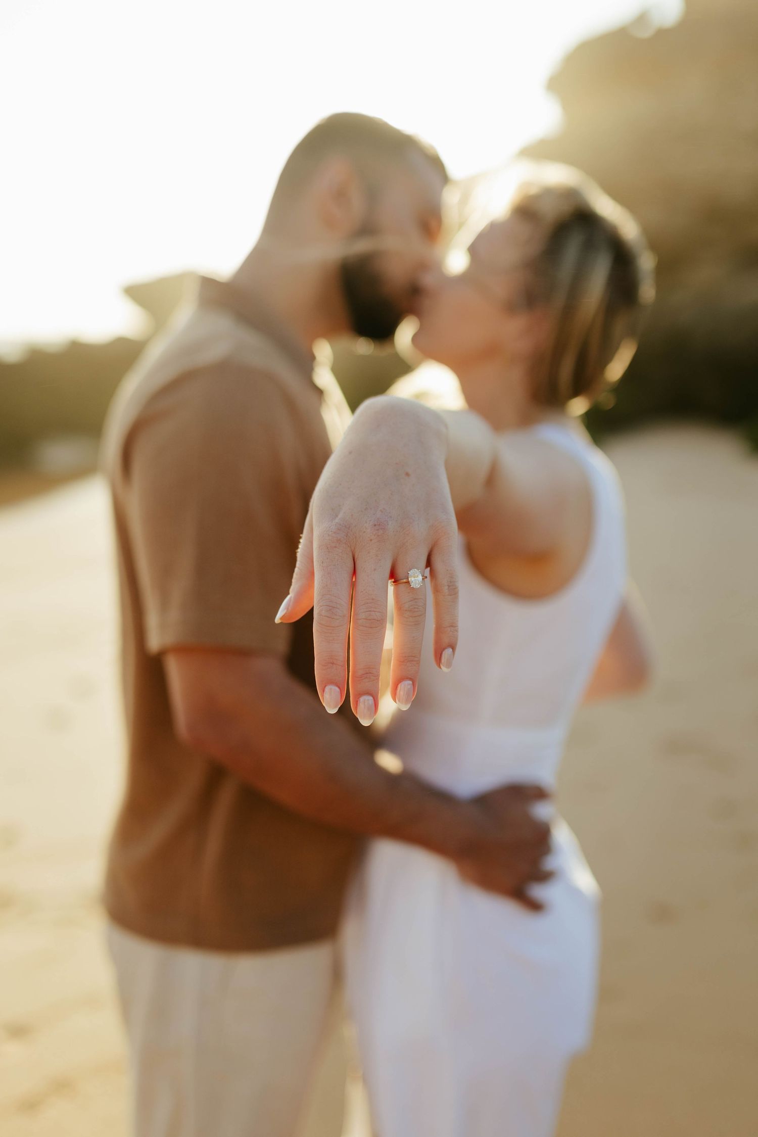 A romantic engagement photo showing a couple kissing at sunset with a diamond ring displayed.