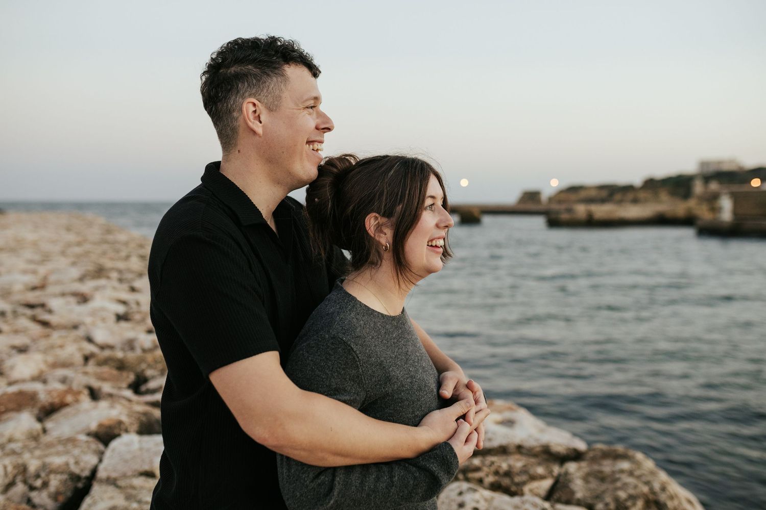 Happy couple embracing on rocky coastline during sunset photo session. Meia Praia, Lagos, Algarve, Portugal.